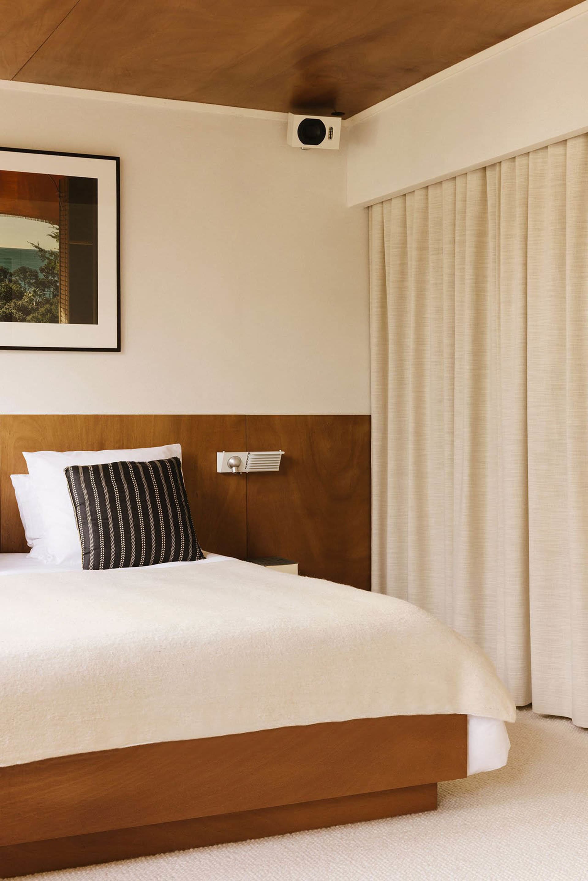 A modern bedroom with a large white bed featuring a black pillow on a wooden headboard, two pictures hanging above, and a bright atmosphere
