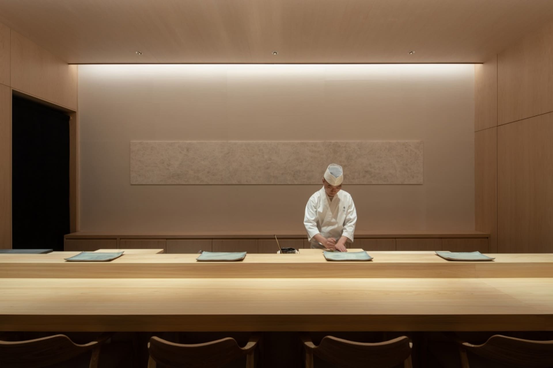 A professional kitchen with a chef preparing food at a wooden dining table, surrounded by chairs and utensils