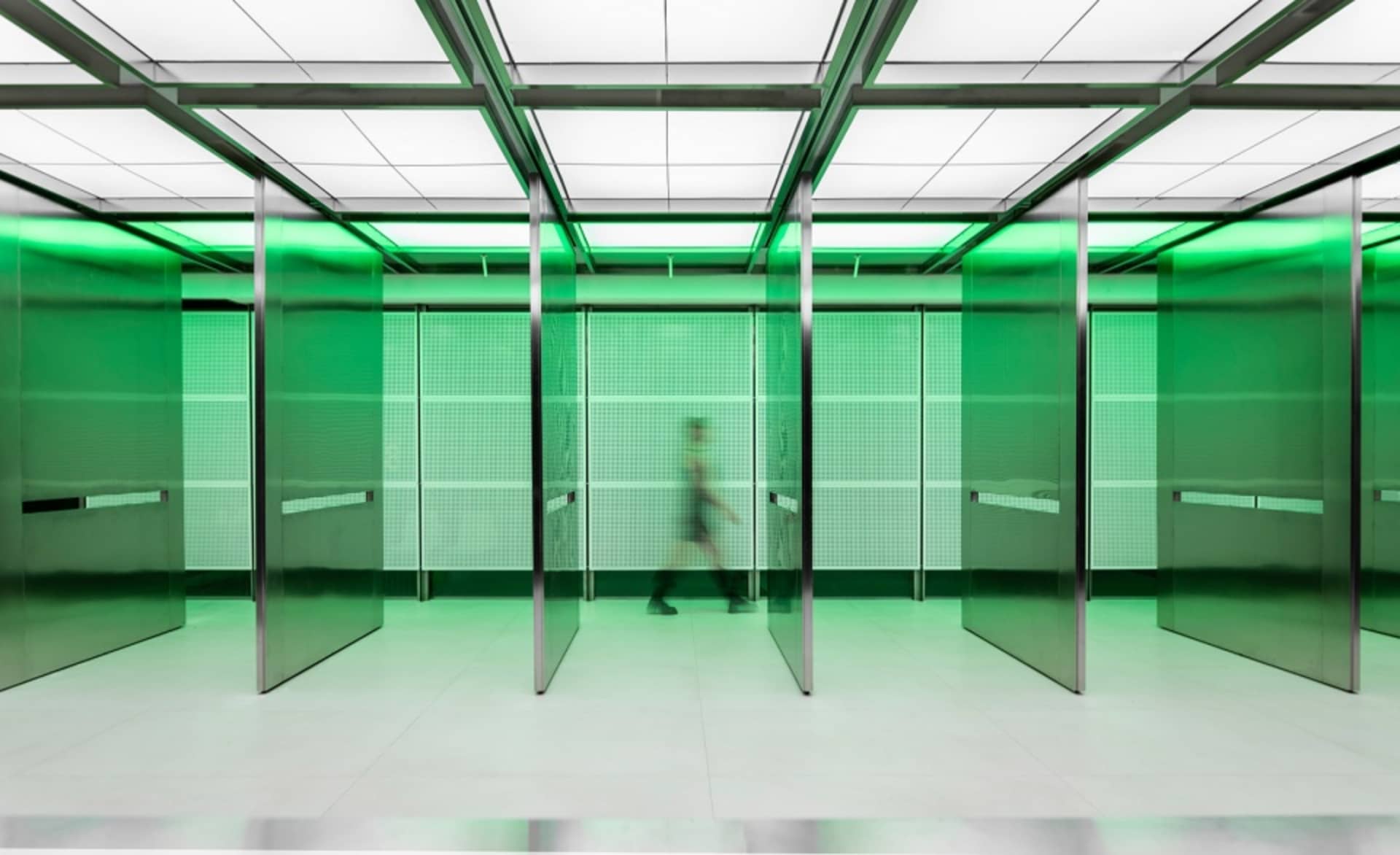 A woman walks down a well-lit office hallway with glass walls, green lighting, and two doors on either side