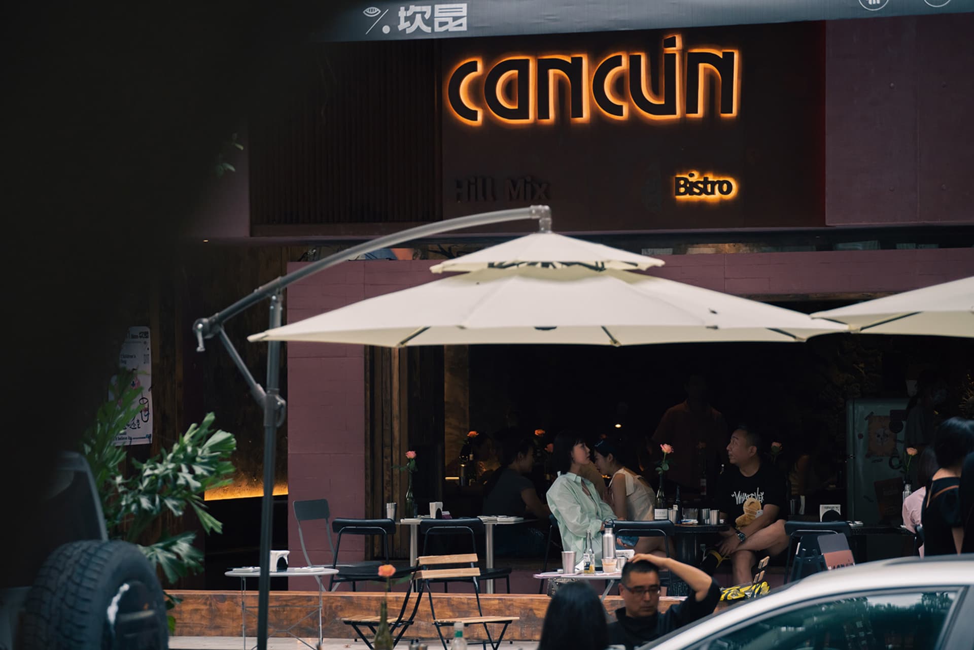 Outdoor dining area with tables, chairs, umbrellas, and potted plants, featuring patrons enjoying their meals in a relaxed atmosphere
