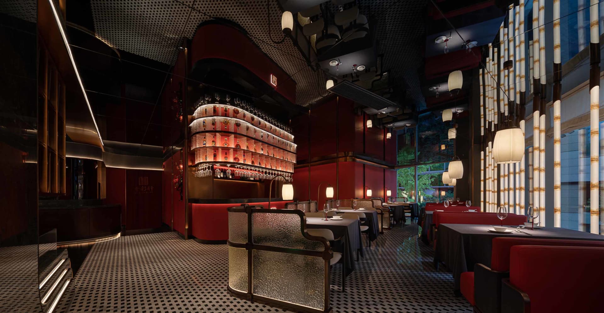 A modern bar area with red walls, a display of liquor bottles, and wine glasses on the countertop, set amidst a lively atmosphere