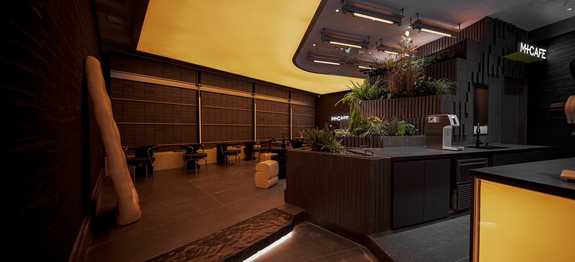 A well-lit interior space with a large yellow wall, multiple chairs, and various potted plants, creating a comfortable atmosphere