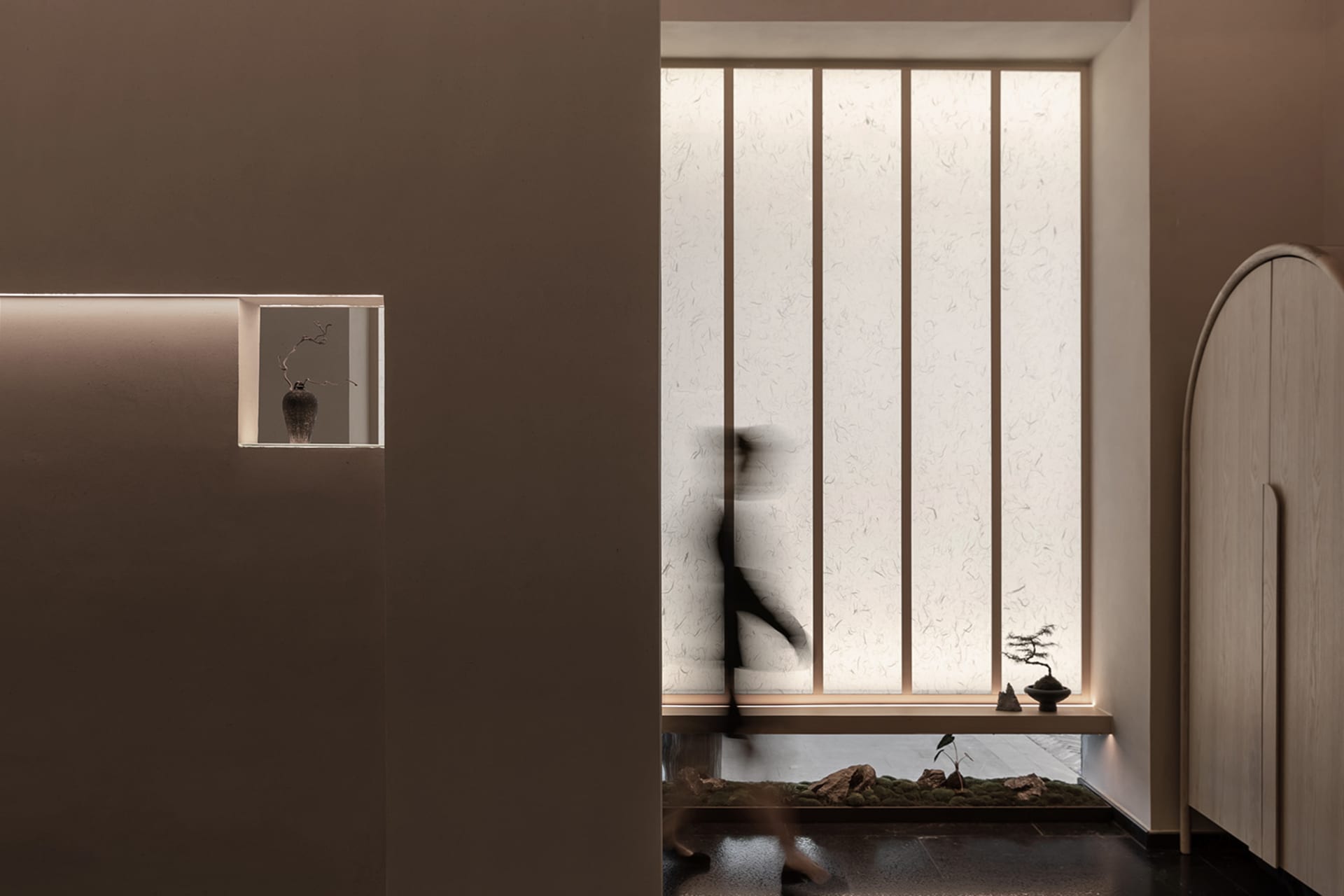 A person walks through a calm and serene interior space with two potted plants and a vase on a surface near the center