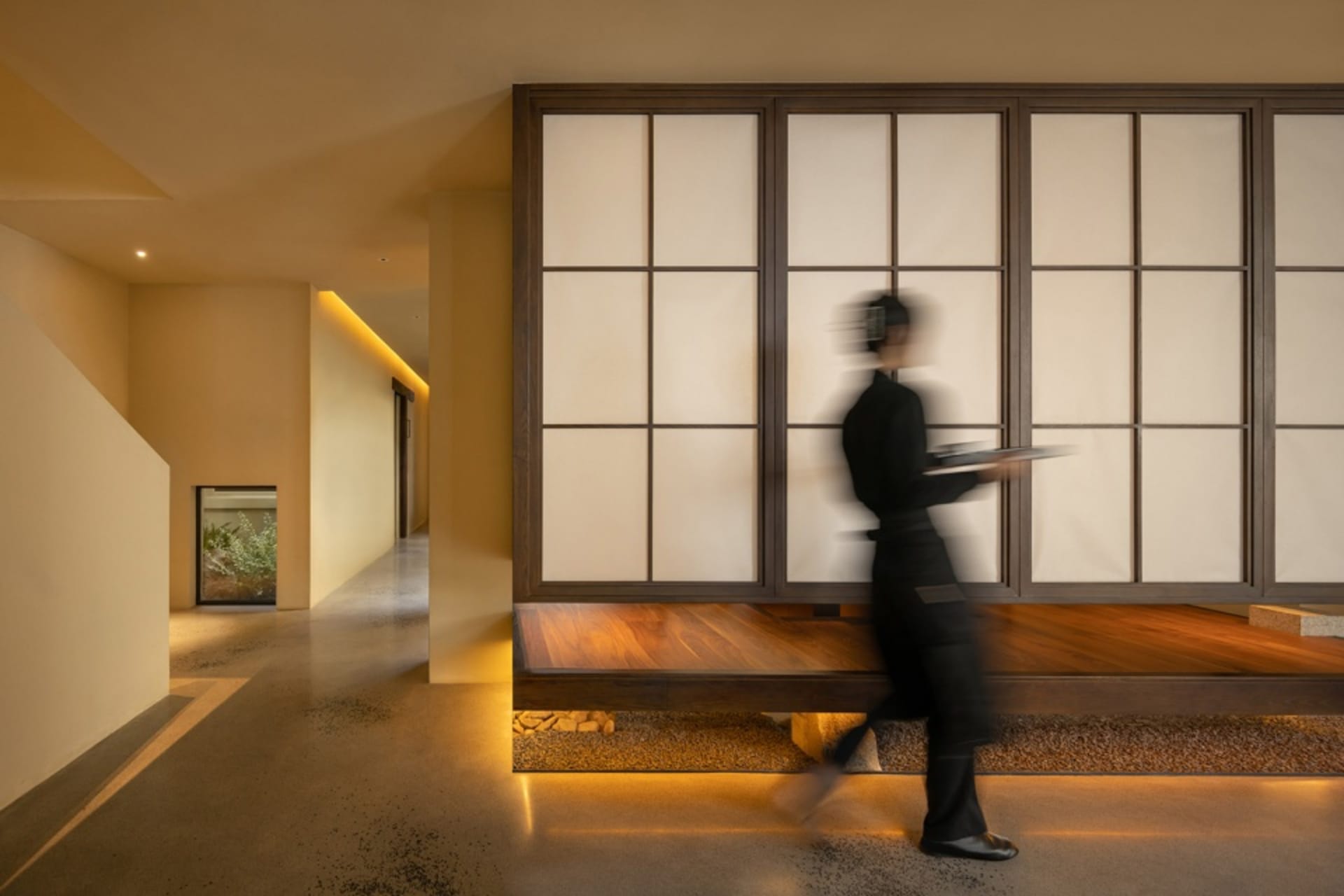 A woman walks through a dimly lit hallway with a wooden bench and a door and window on either side