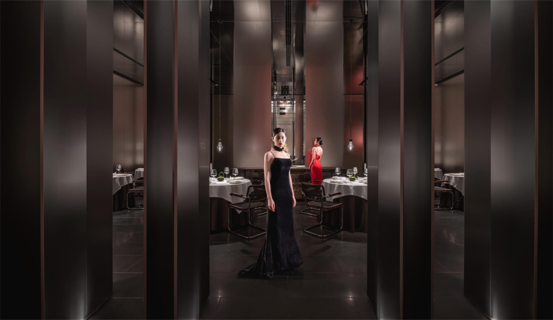 A woman in a black dress stands in front of a dining table with wine glasses and chairs in a sophisticated restaurant setting