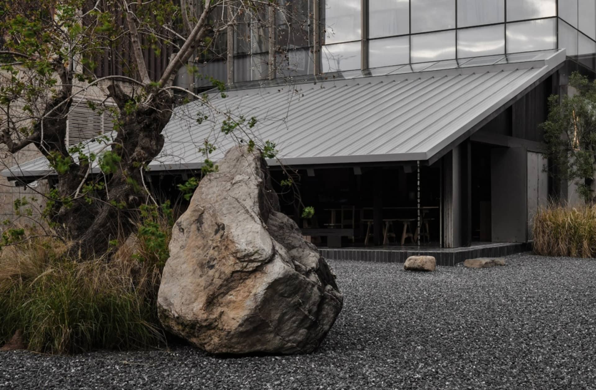 A building with a gray roof and a large rock in front, surrounded by outdoor seating and natural surroundings