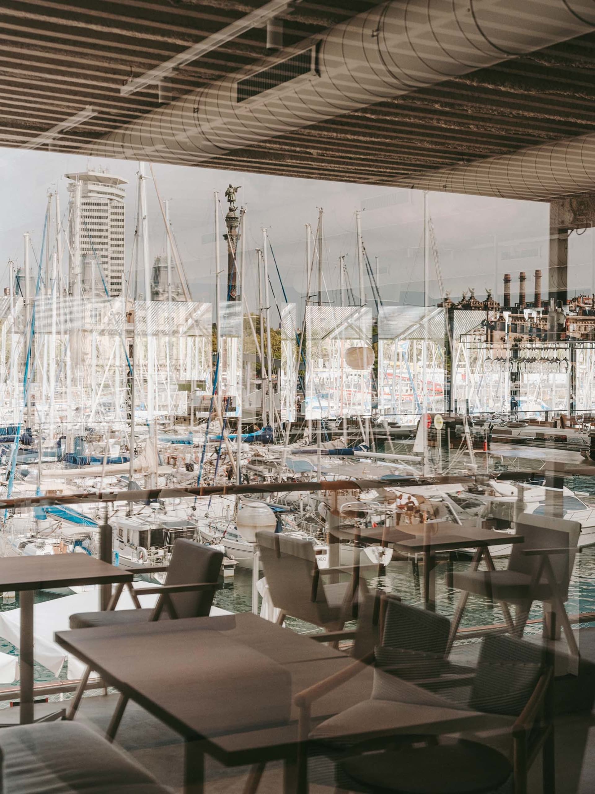 A restaurant interior with multiple dining tables and chairs, surrounded by glass windows showing boats, in a well-lit space with a nautical theme