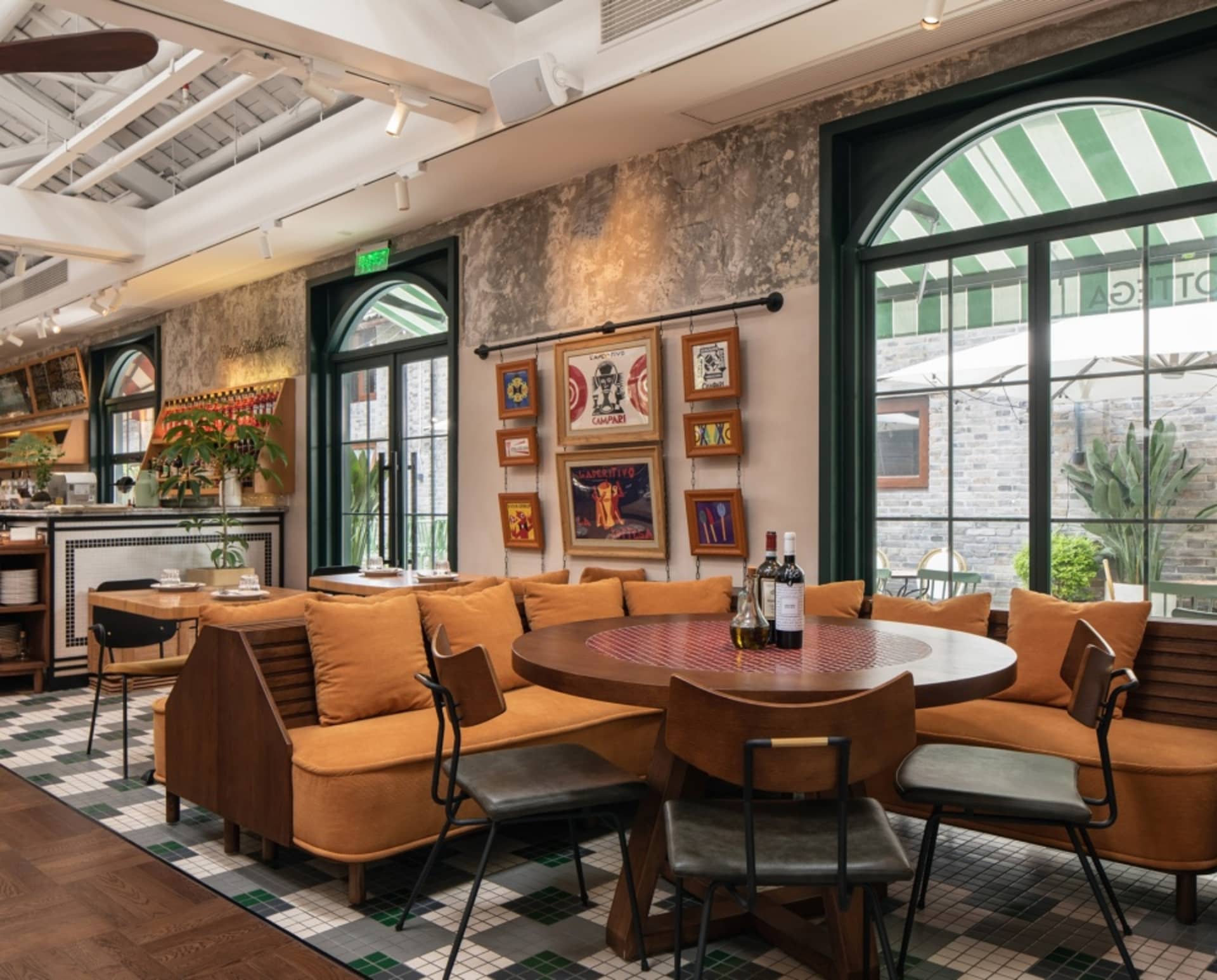 Cozy restaurant dining area with multiple couches and chairs around a large round table, wine bottles on the table, and a warm atmosphere