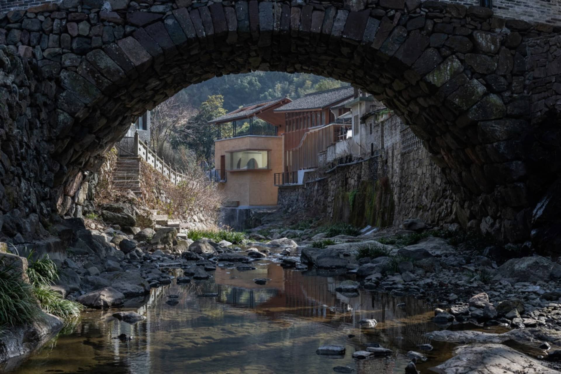 A stone bridge with an archway leads to a brick building in a historic European setting with water nearby