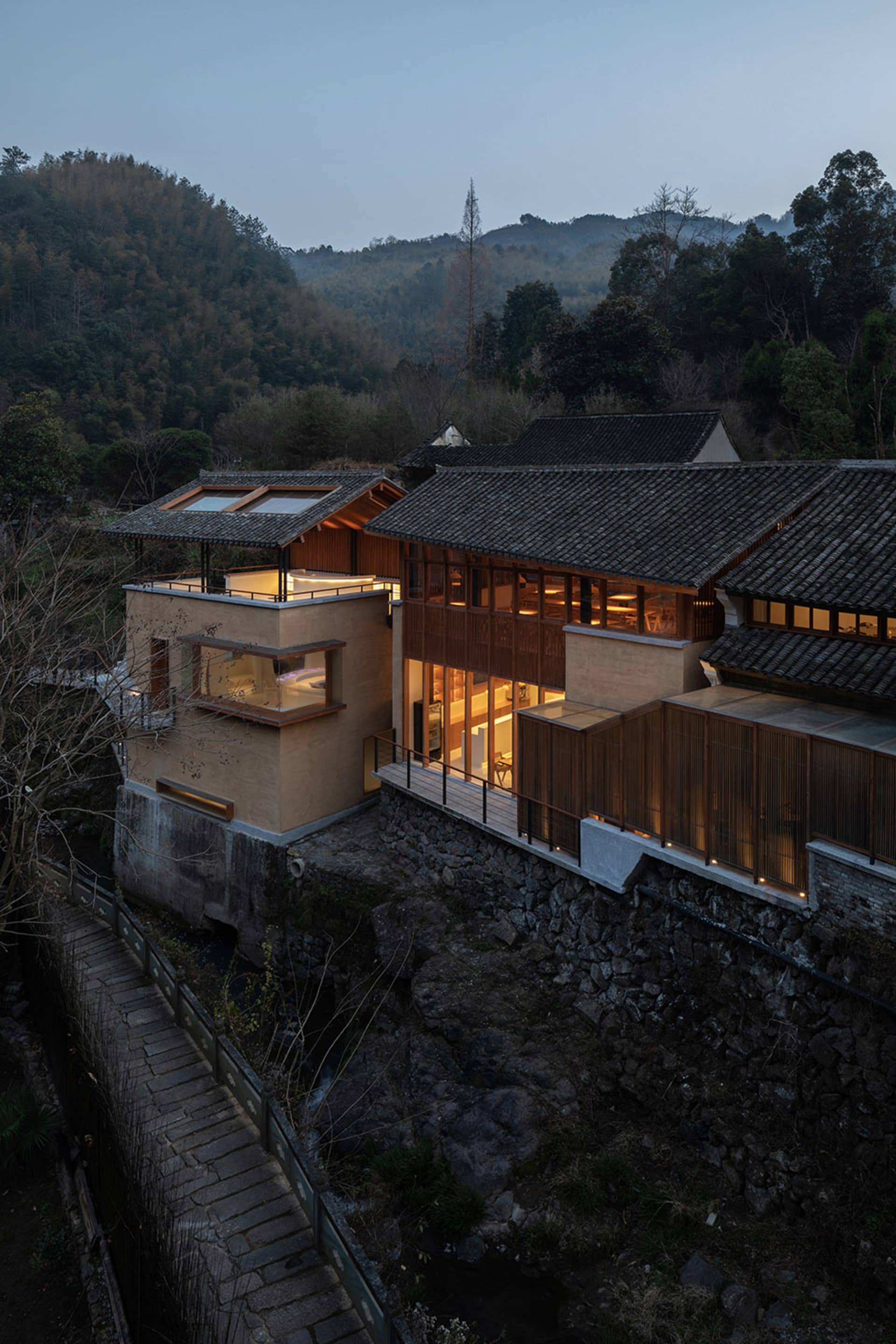 Asian-style house with a wooden exterior, situated on a hillside overlooking a river, with a large open area leading to a patio and several windows allowing natural light