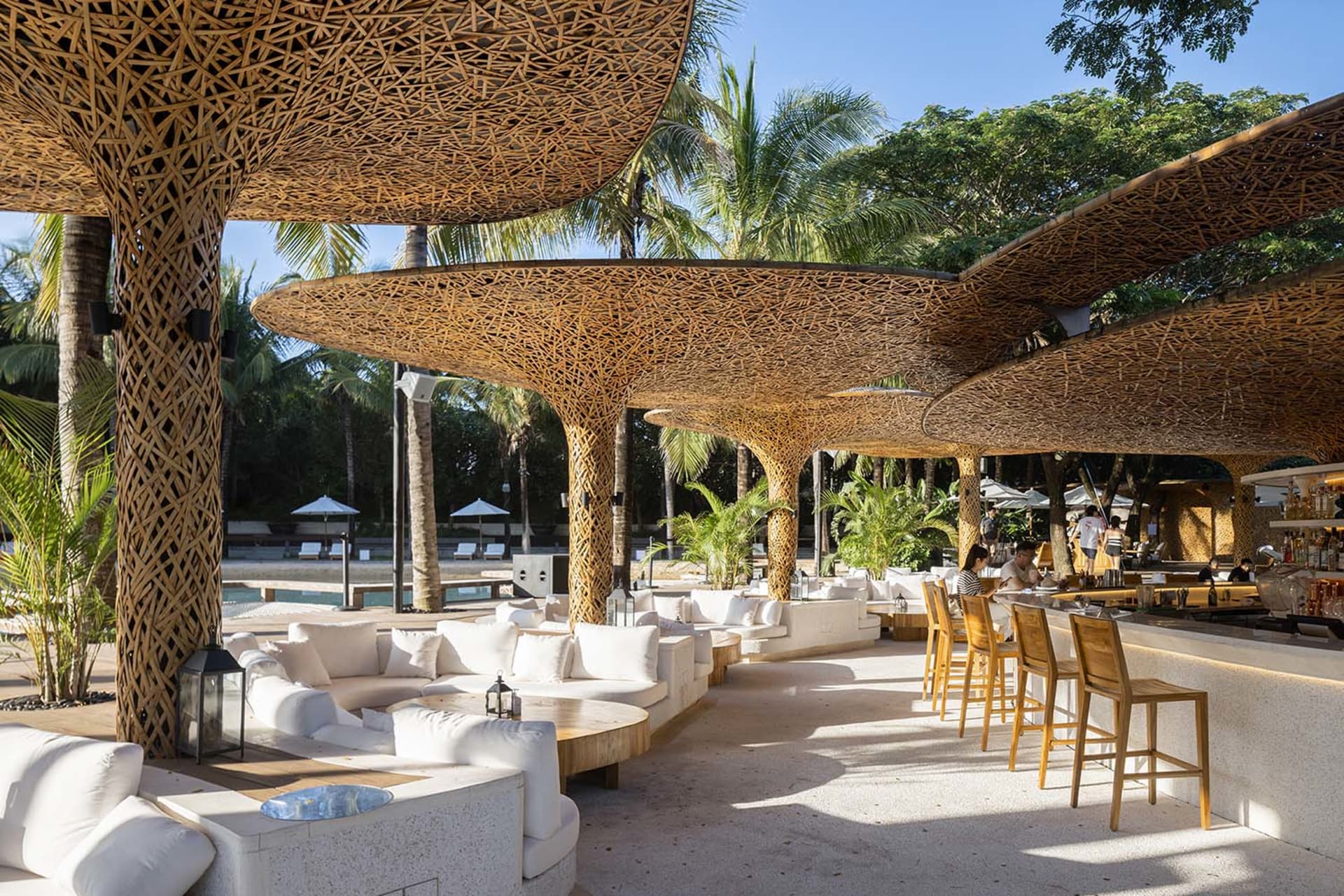 A relaxed outdoor dining space with several tables and chairs under large wooden umbrellas, surrounded by a warm and inviting atmosphere