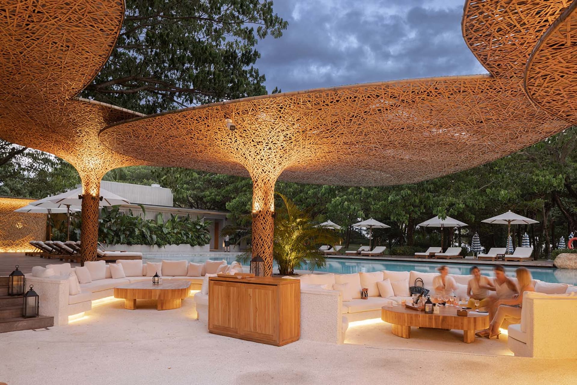 A resort patio with a wooden bar, lounge chairs, dining tables, umbrellas, and a swimming pool, set in a relaxing and inviting atmosphere
