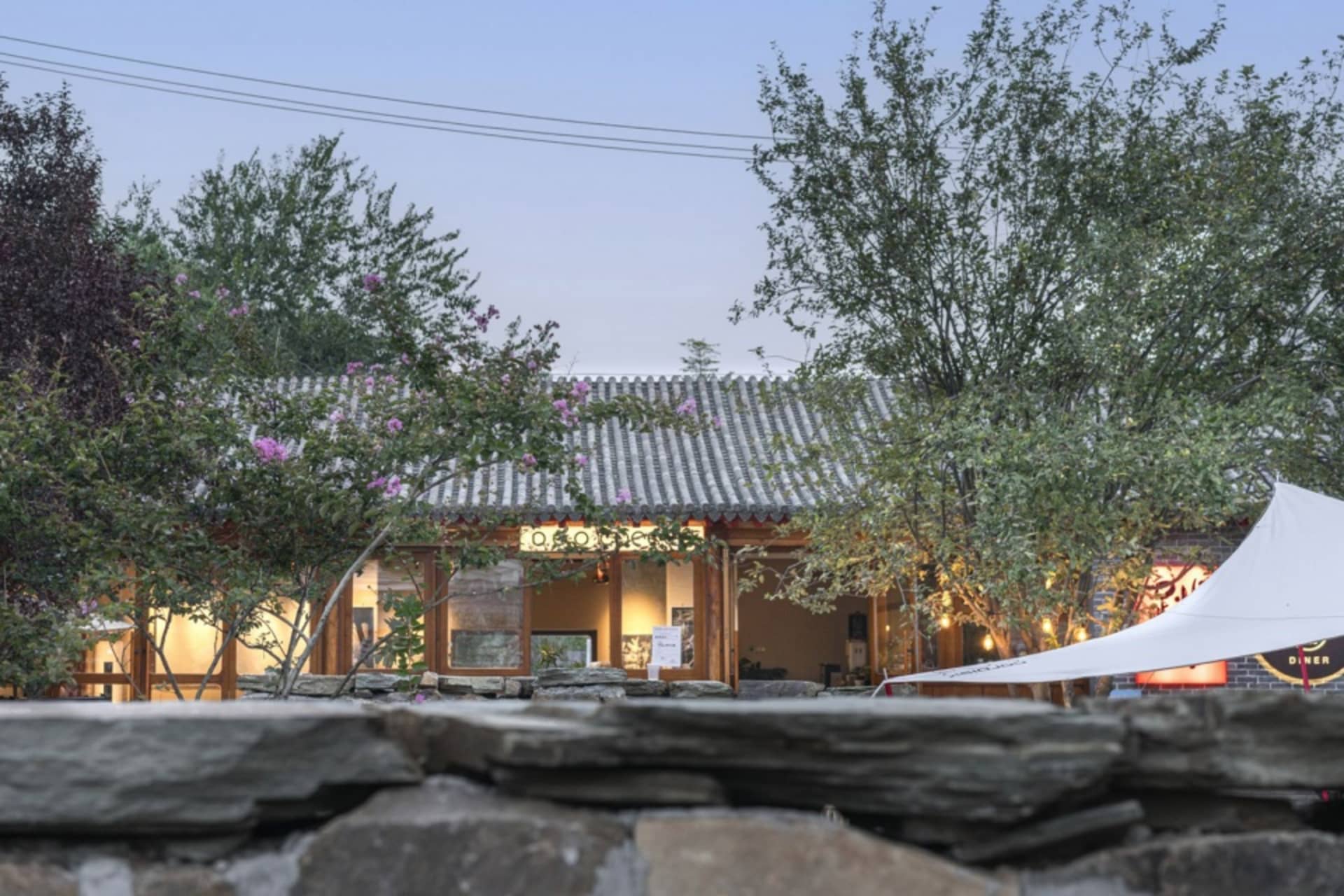 A serene photo of a traditional Asian building with a stone roof, surrounded by trees, flowers, and potted plants, featuring a combination of stone and wood elements, with two umbrellas adding shade or decoration