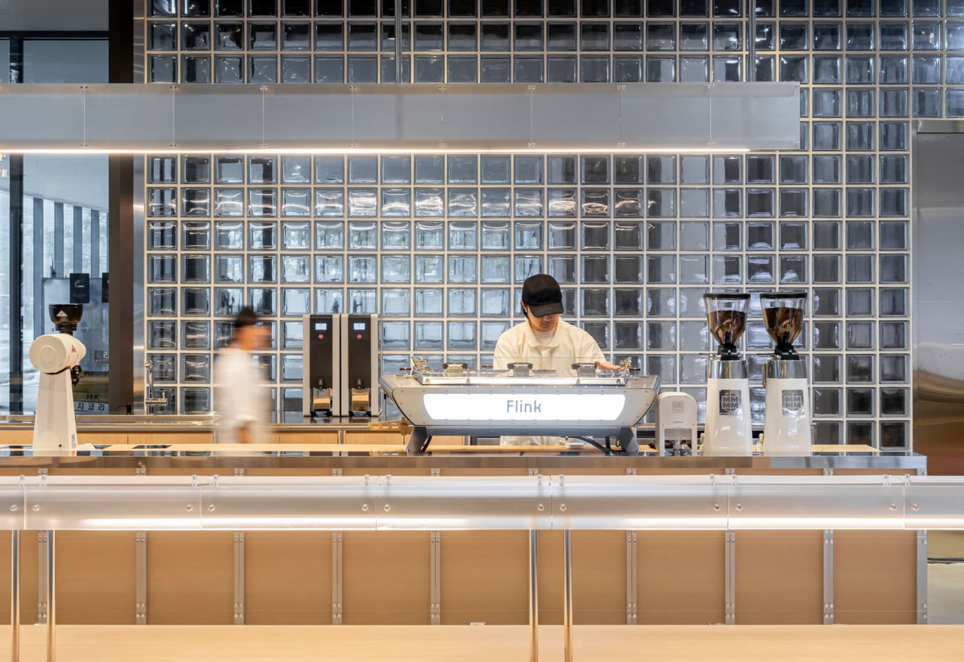 A modern coffee shop with a barista preparing drinks at a counter, surrounded by warm wood tones and metal accents, with two customers in the scene