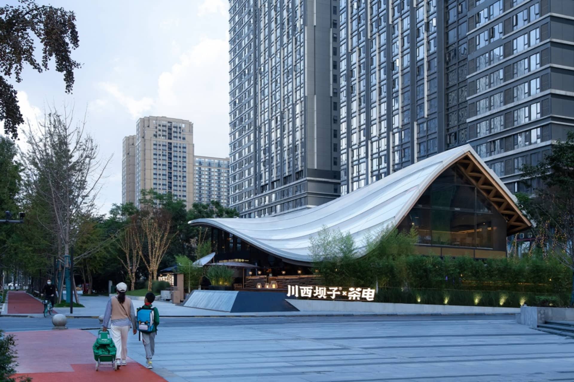 A bustling city street scene with a large building featuring a curved roof, people walking on the sidewalk, and a stroller being pushed by two women