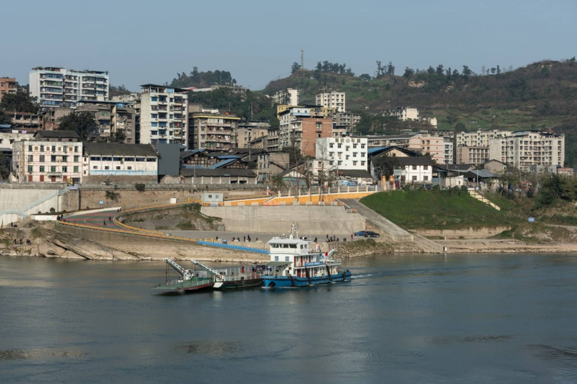 A bustling riverfront scene with a large boat and brick buildings, surrounded by people going about their daily activities