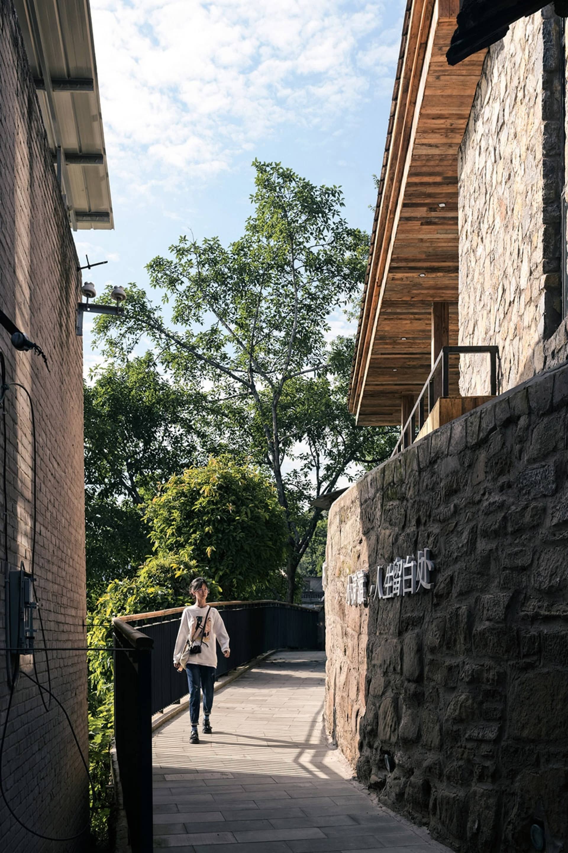 An Asian woman walks down a stone pathway in a courtyard with a mix of stone and wood architecture, surrounded by trees and a sense of tranquility