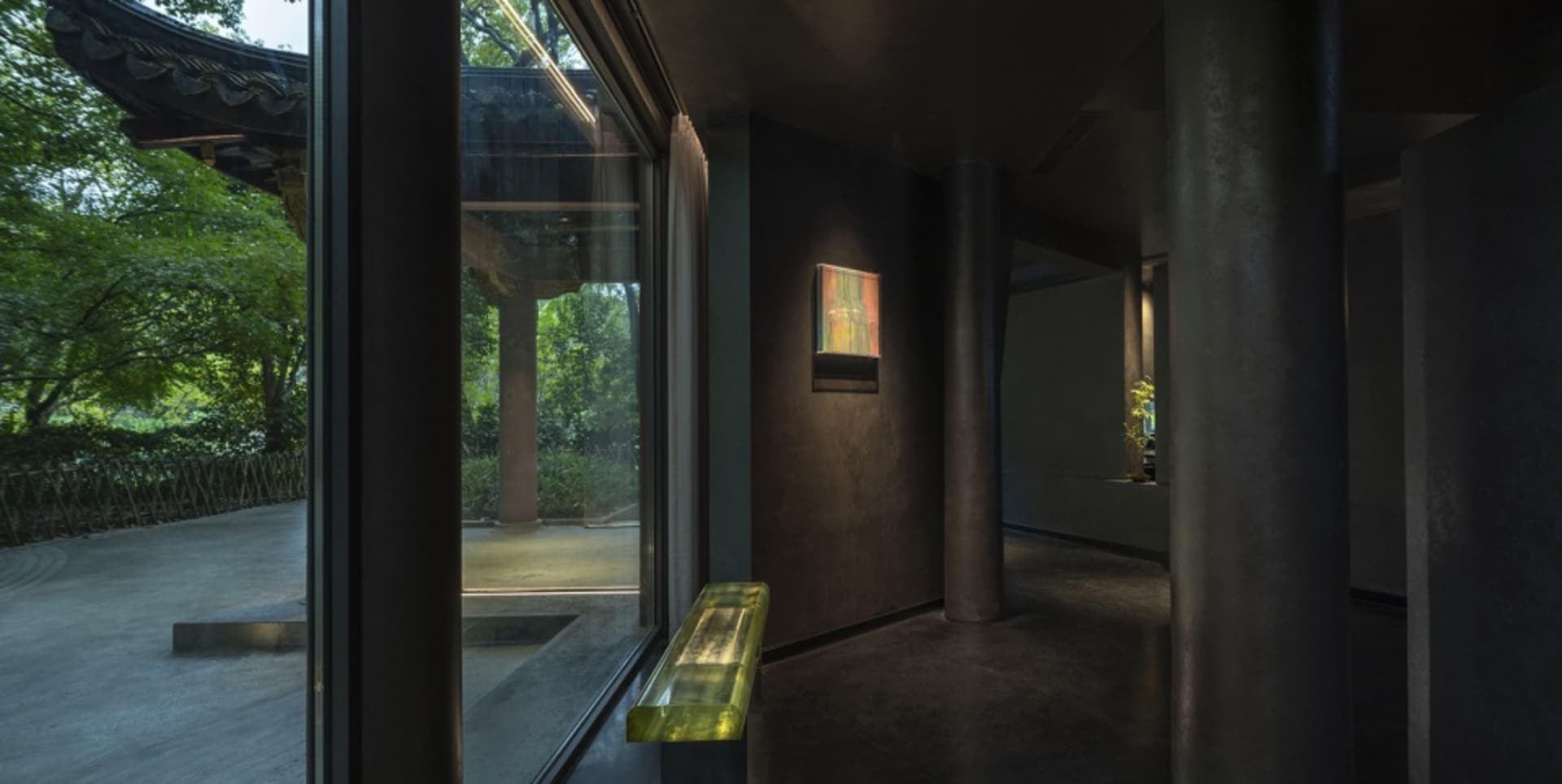 Dimly lit room with a bench near a window, two potted plants, and a painting on display