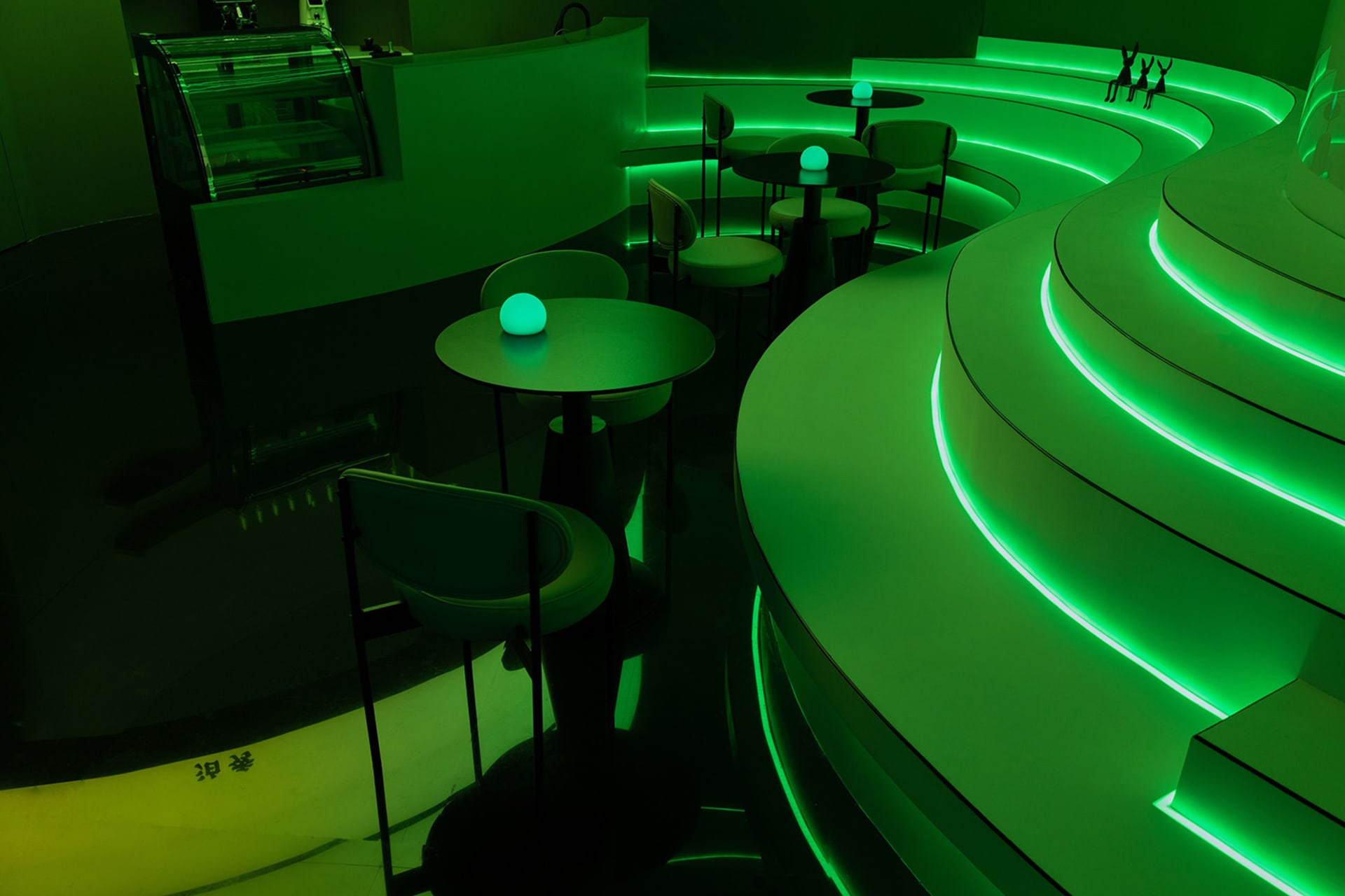 A modern bar area with green neon lights on the walls and tables, surrounded by chairs and well-lit by large windows or other light sources