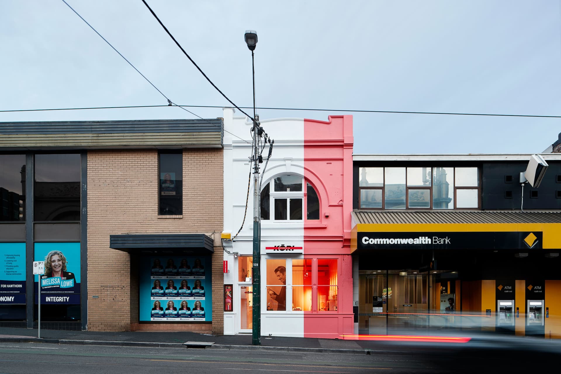 Two adjacent buildings with red and white stripes and white facades, illuminated storefronts, and people walking outside, set against a city street with parked cars