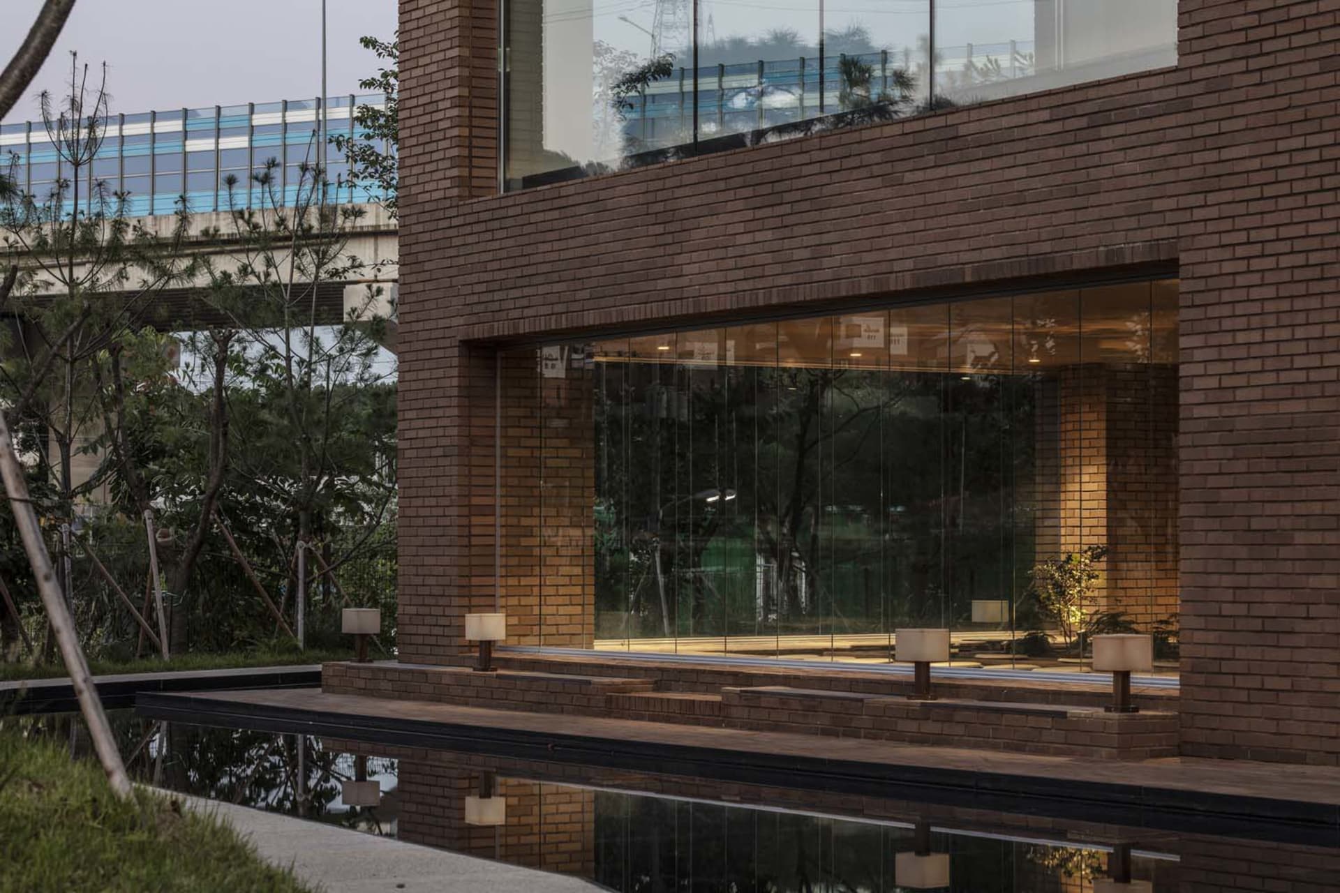 A building with a brick exterior and a large glass wall overlooking a pool area with potted plants, near a train line