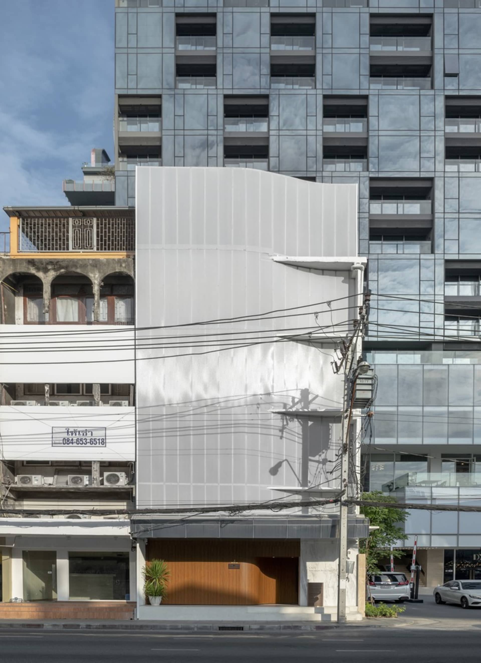 A city street view of a tall building with a white concrete facade, featuring windows on various levels and greenery on the street