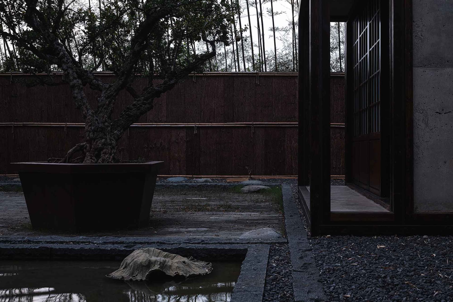A serene Asian-style garden with a pond, wooden building, and stone walkway, surrounded by trees and a peaceful atmosphere