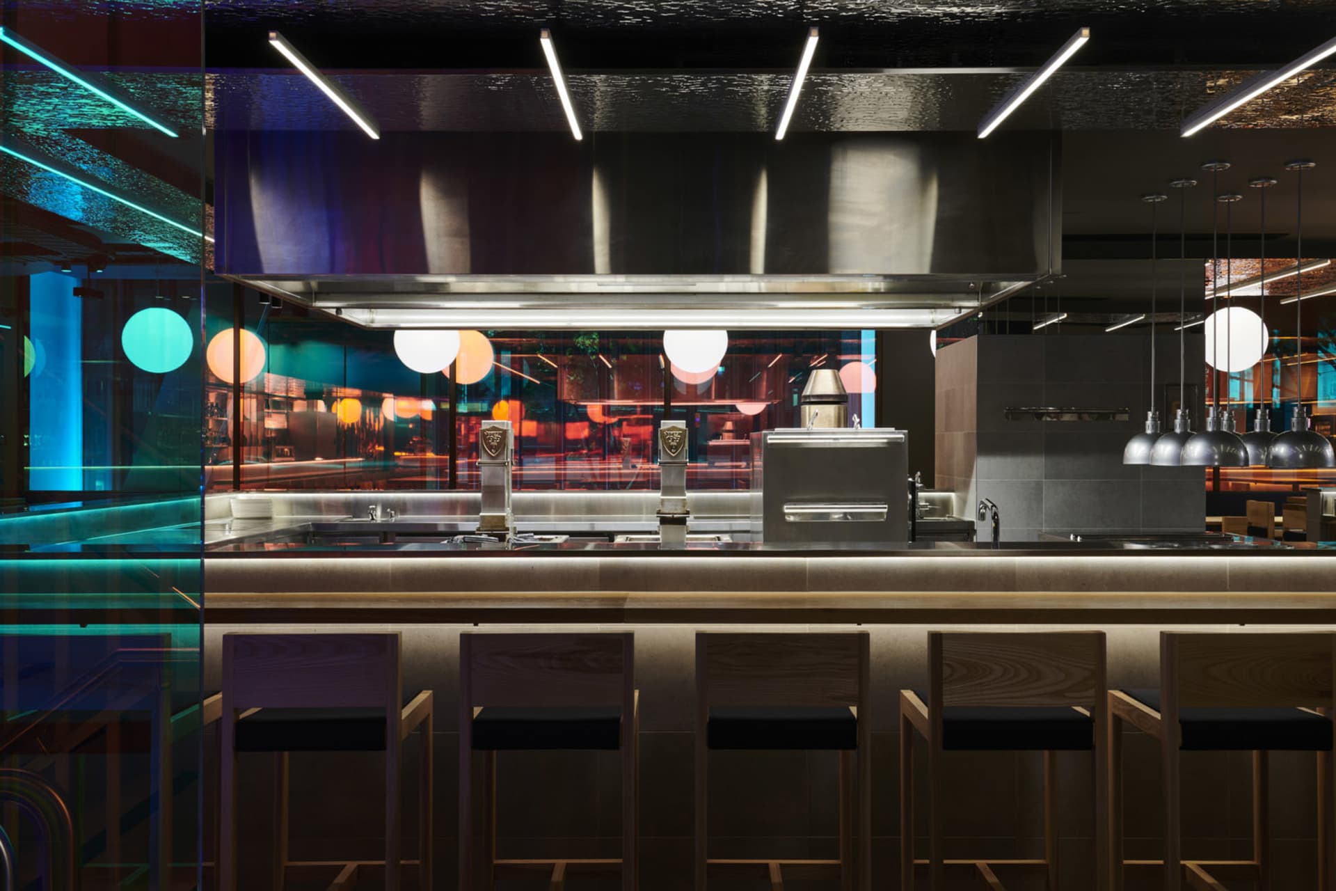 Industrial kitchen with stainless steel appliances and counters, featuring a refrigerator, oven, and sink, with barstools and cups on the counter