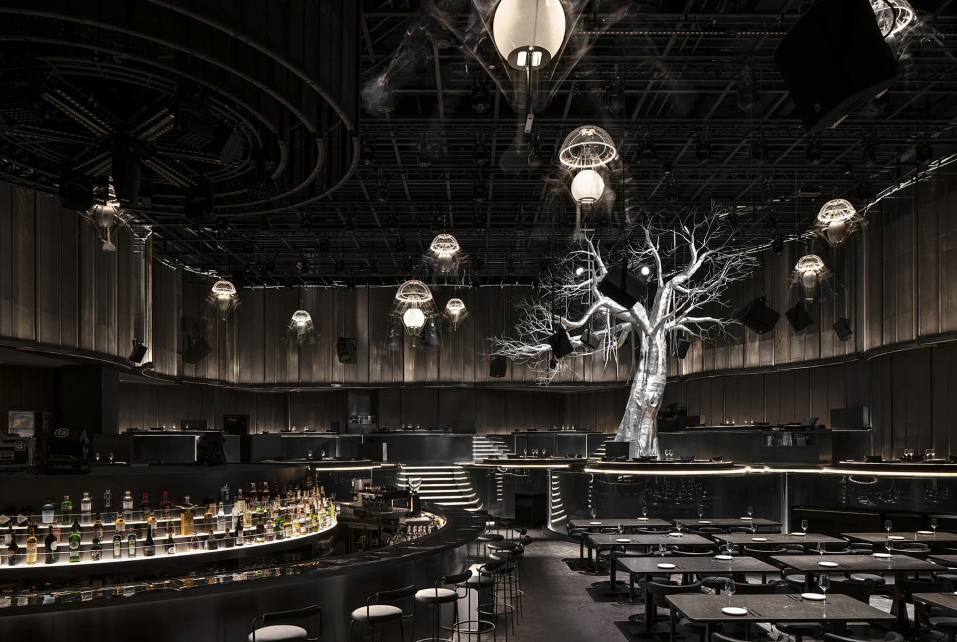 A warm and inviting bar area with a tree sculpture centerpiece, surrounded by tables and chairs, and bottles on surfaces