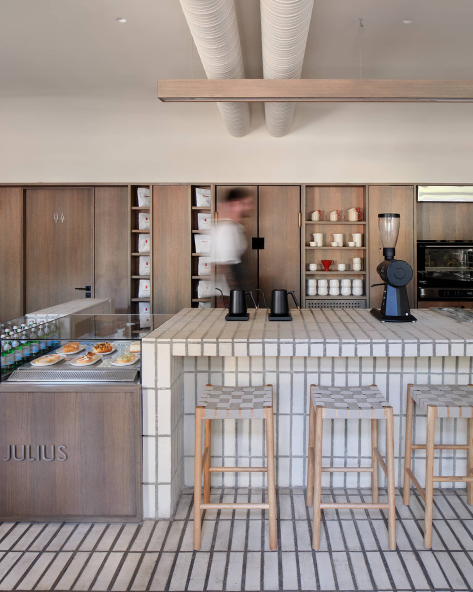 A restaurant kitchen with wooden cabinets, tile flooring, and a bar with stools, featuring various items on the countertops and a microwave