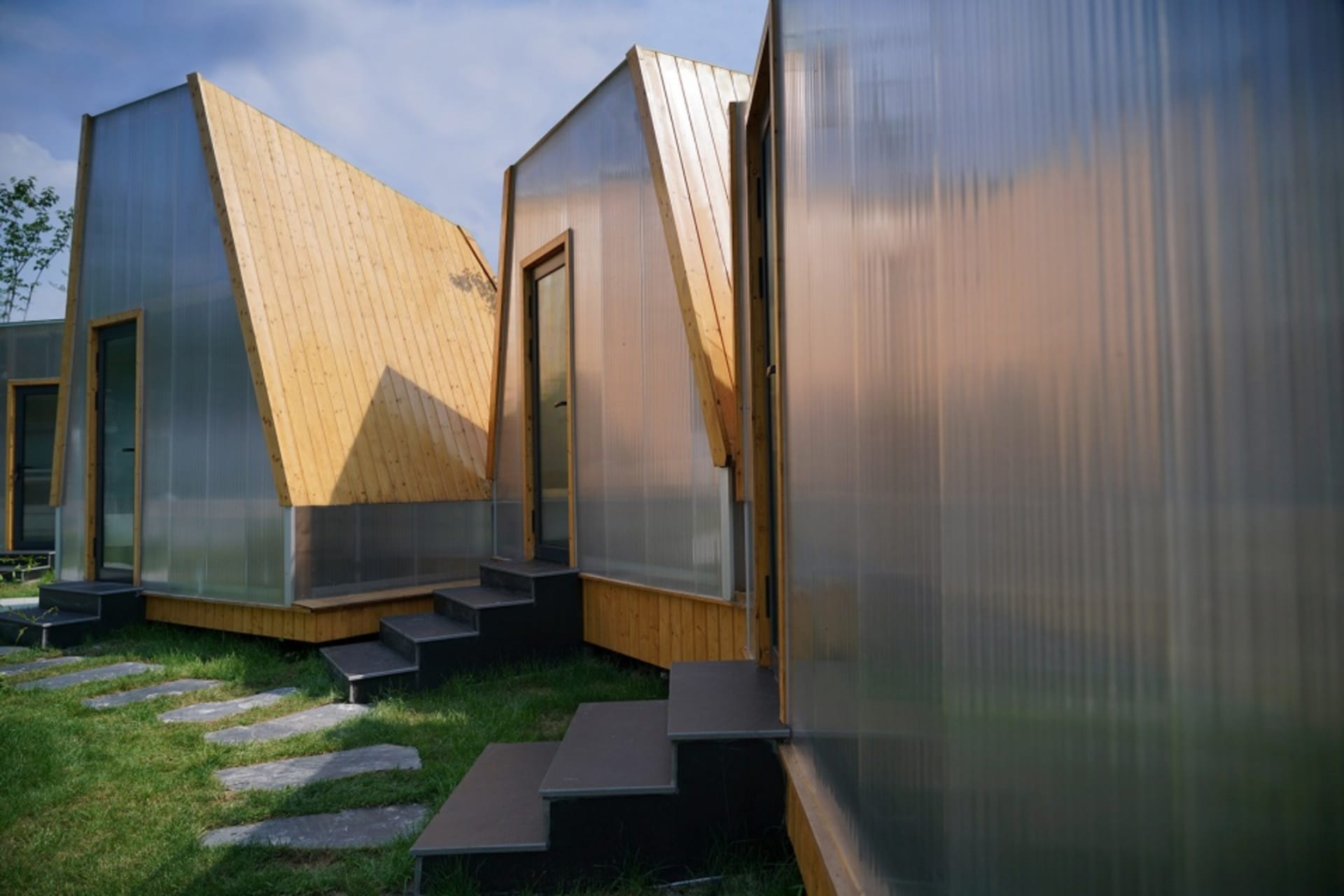 Two modern houses with glass walls and wooden frames, situated on a grassy area with stairs leading up to their entrances