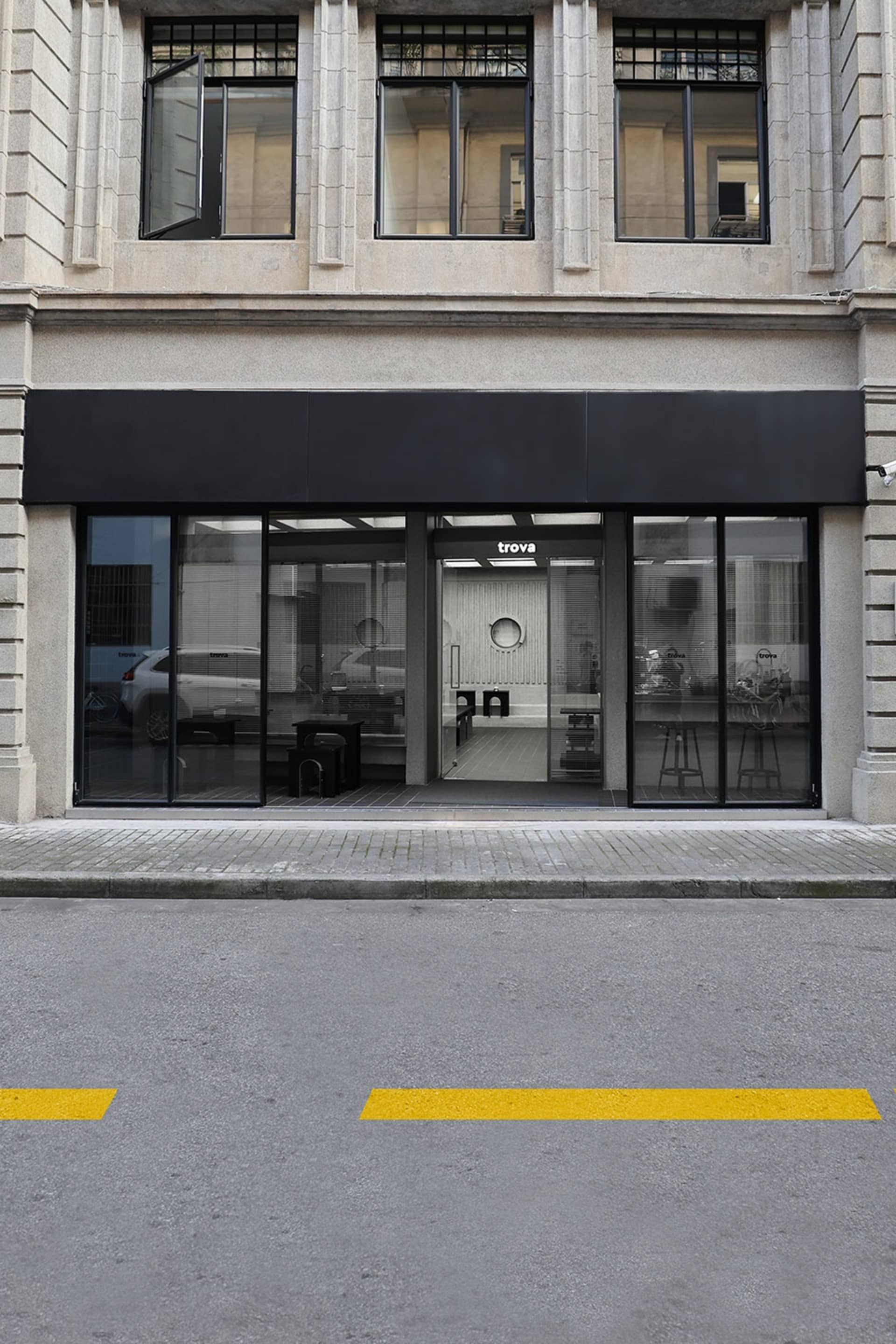 A city storefront with a black awning, large window, and potted plants outside, set against a backdrop of urban surroundings