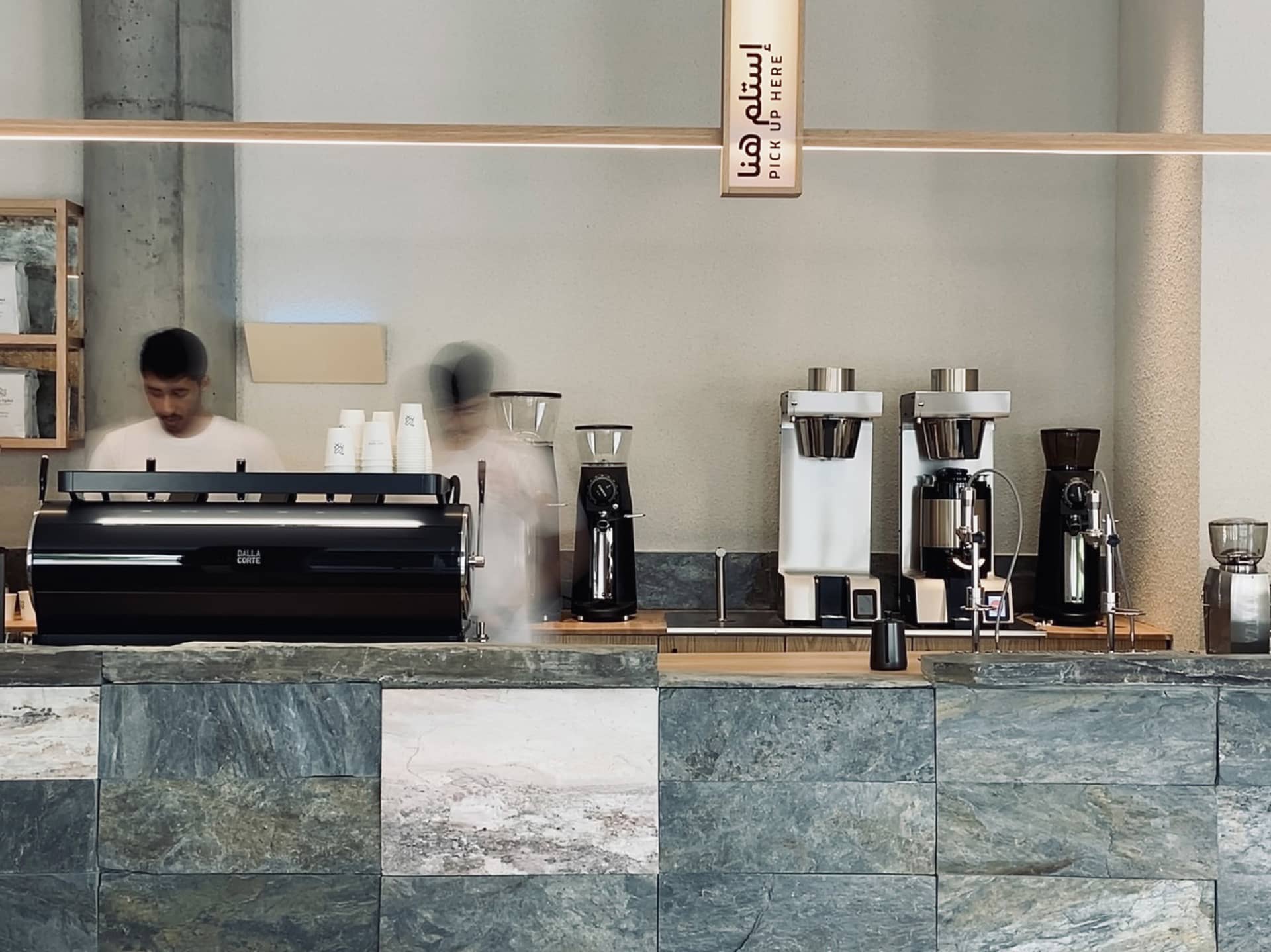 A casual coffee shop interior with a counter featuring multiple coffee machines and cups, and two people in the background