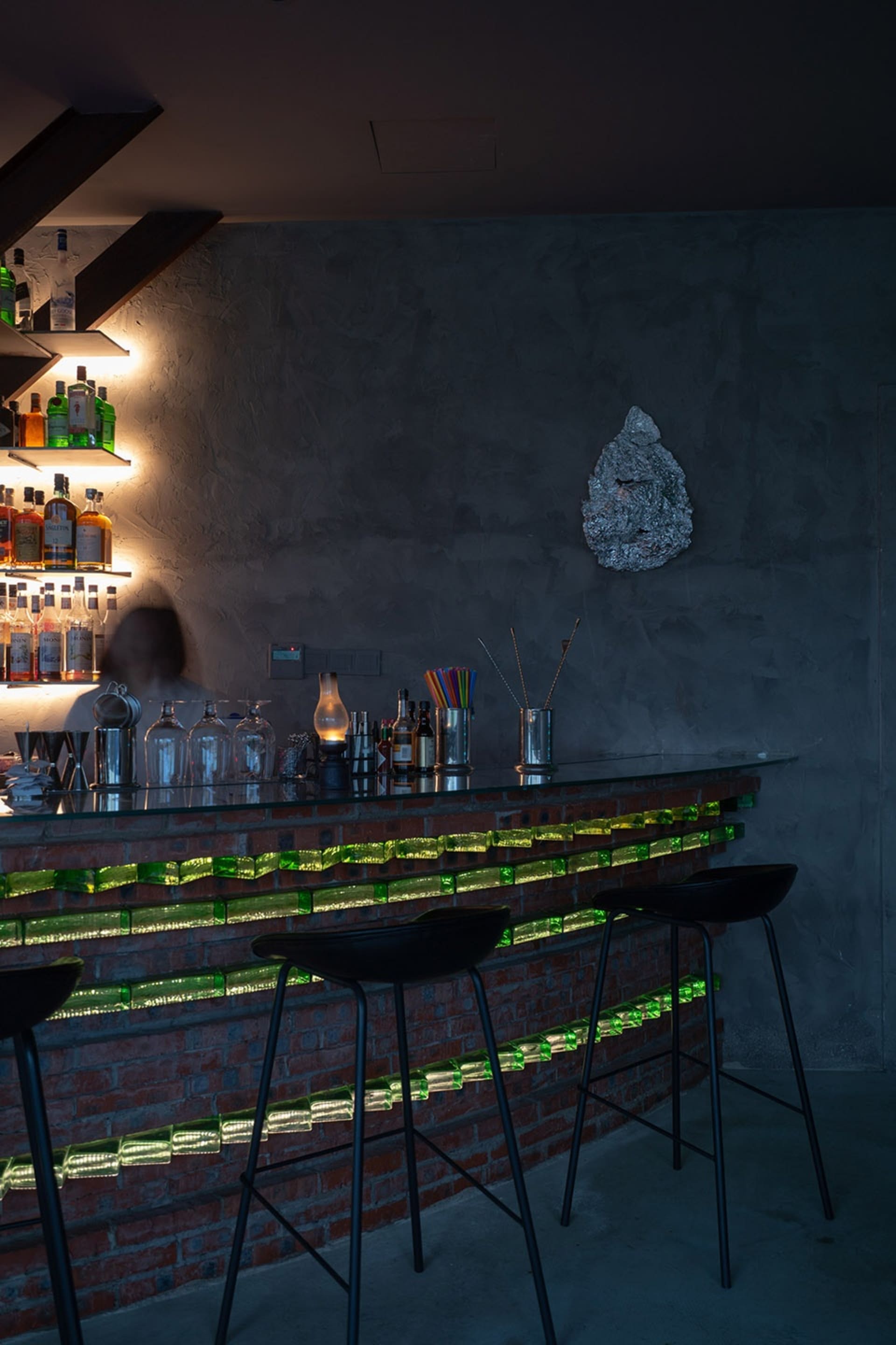 Modern bar area with a green backsplash, brick wall, glass front, and stools, featuring bottles, a vase, and a bowl