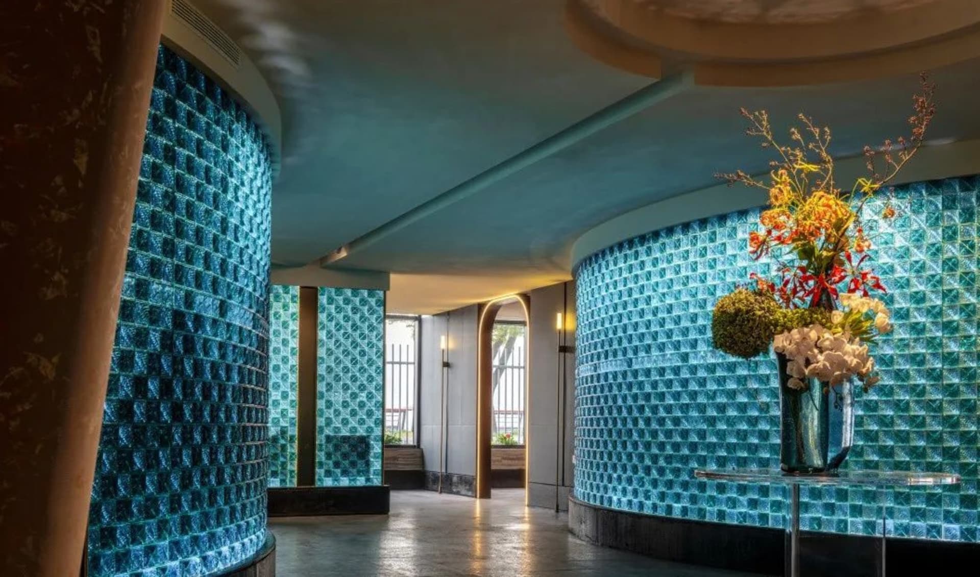 A calm and inviting hallway with blue walls, a glass block ceiling, and potted plants along the length of the space