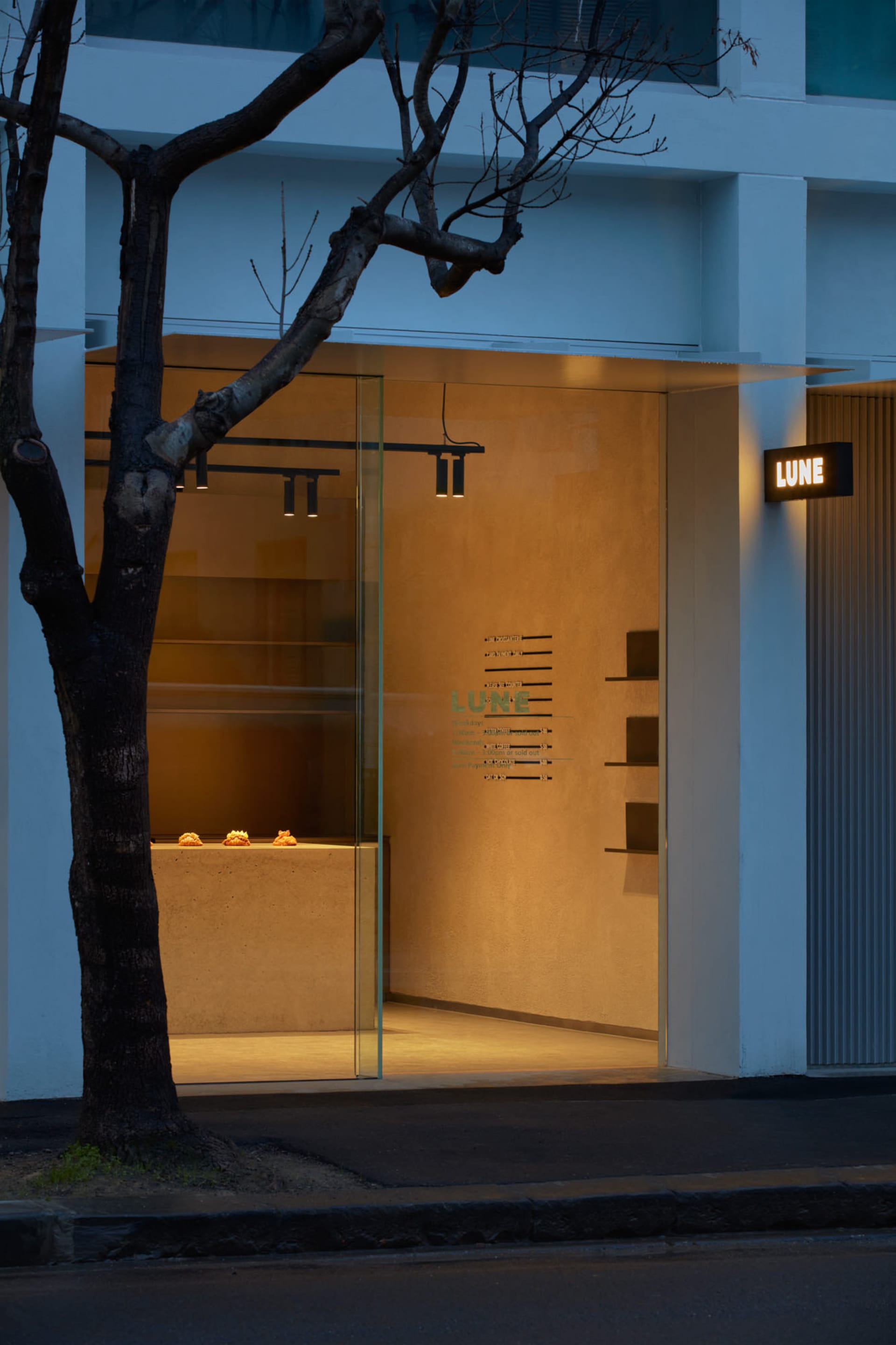 A brightly lit storefront with a glass door, surrounded by two trees and featuring three cakes on display