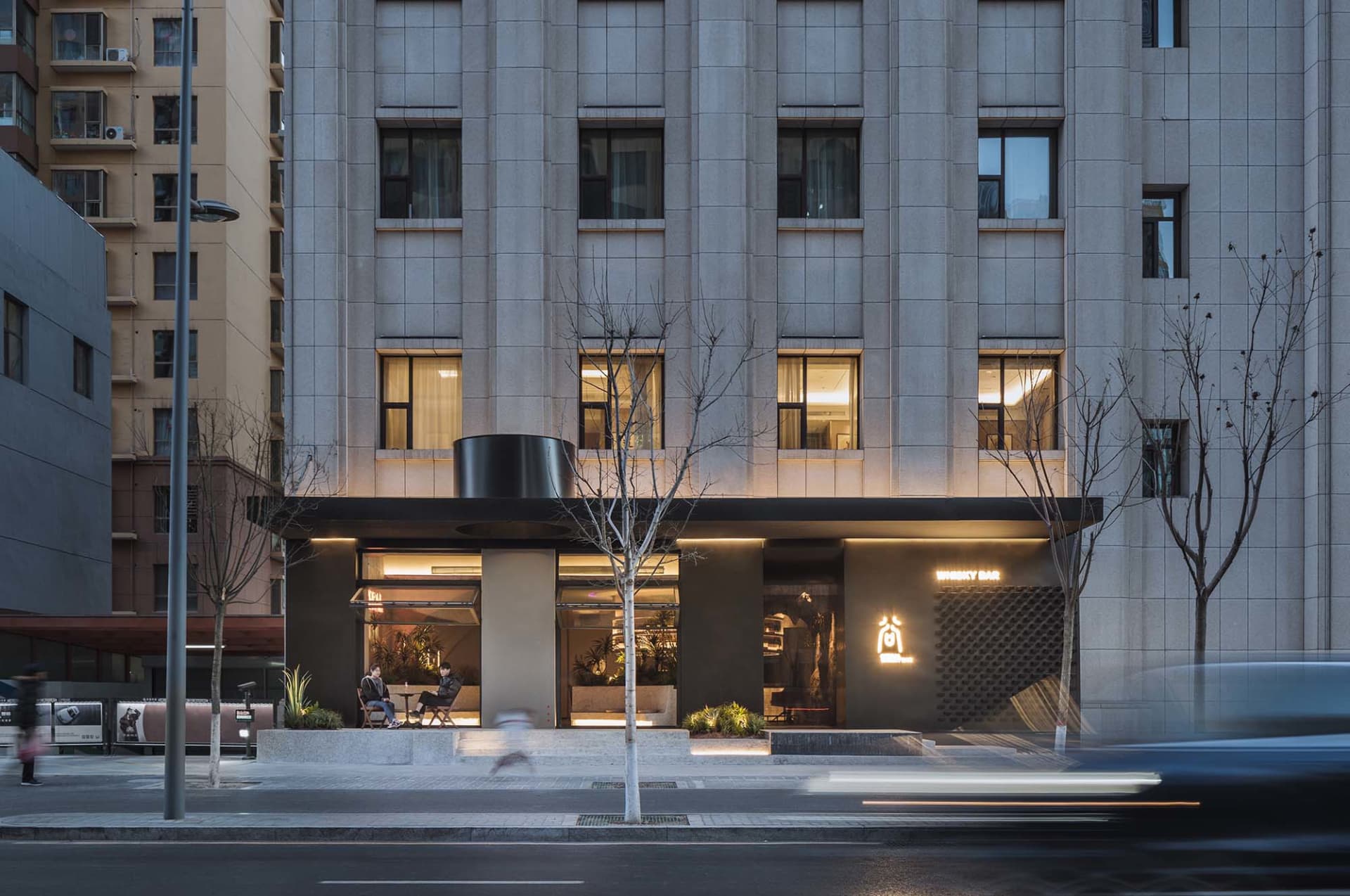 Dark modern building facade at night with a tree in front, featuring windows with interior lights and two outdoor chairs