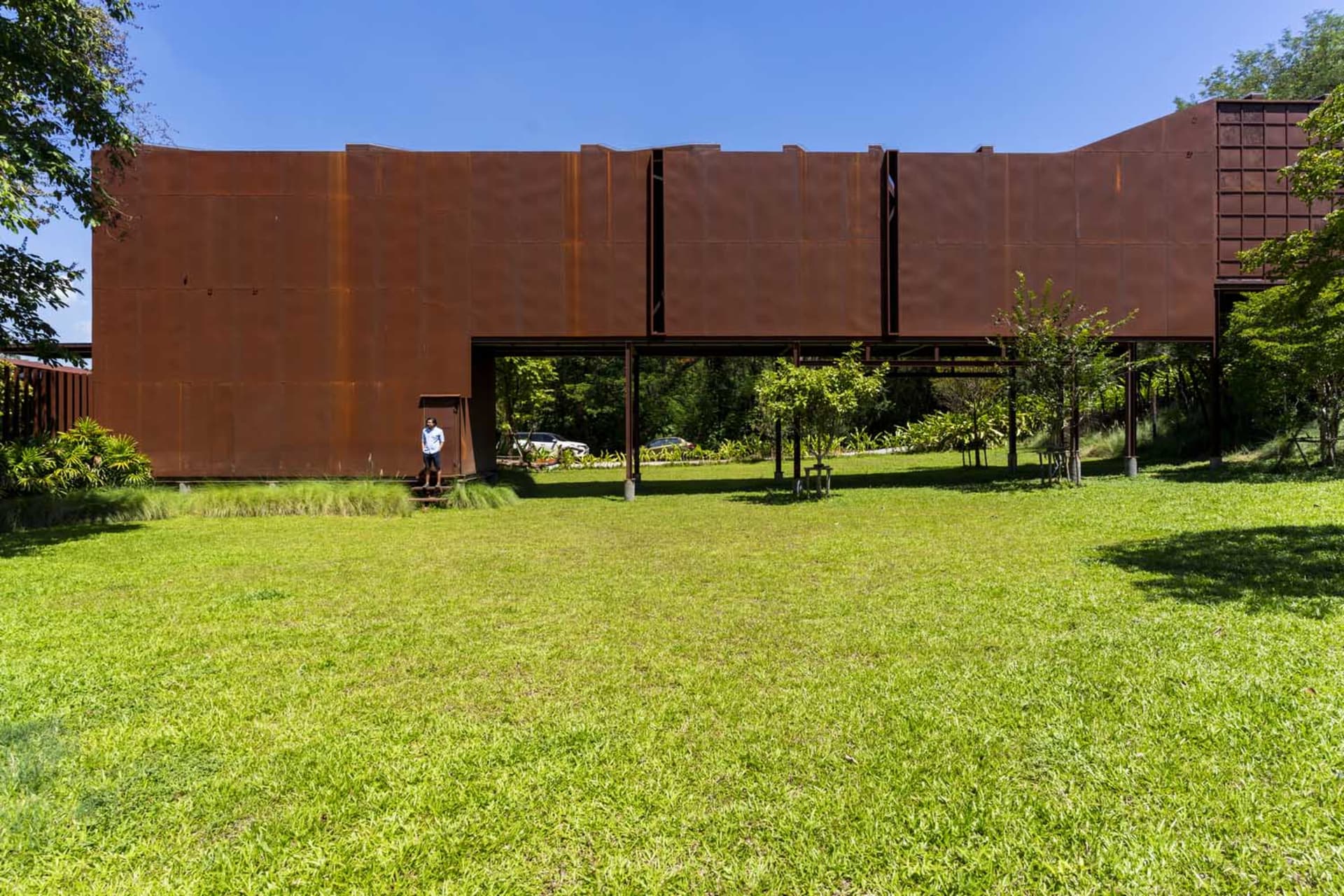 A relaxed outdoor space with a metal structure in the background, benches scattered throughout, and lush greenery adding to the ambiance