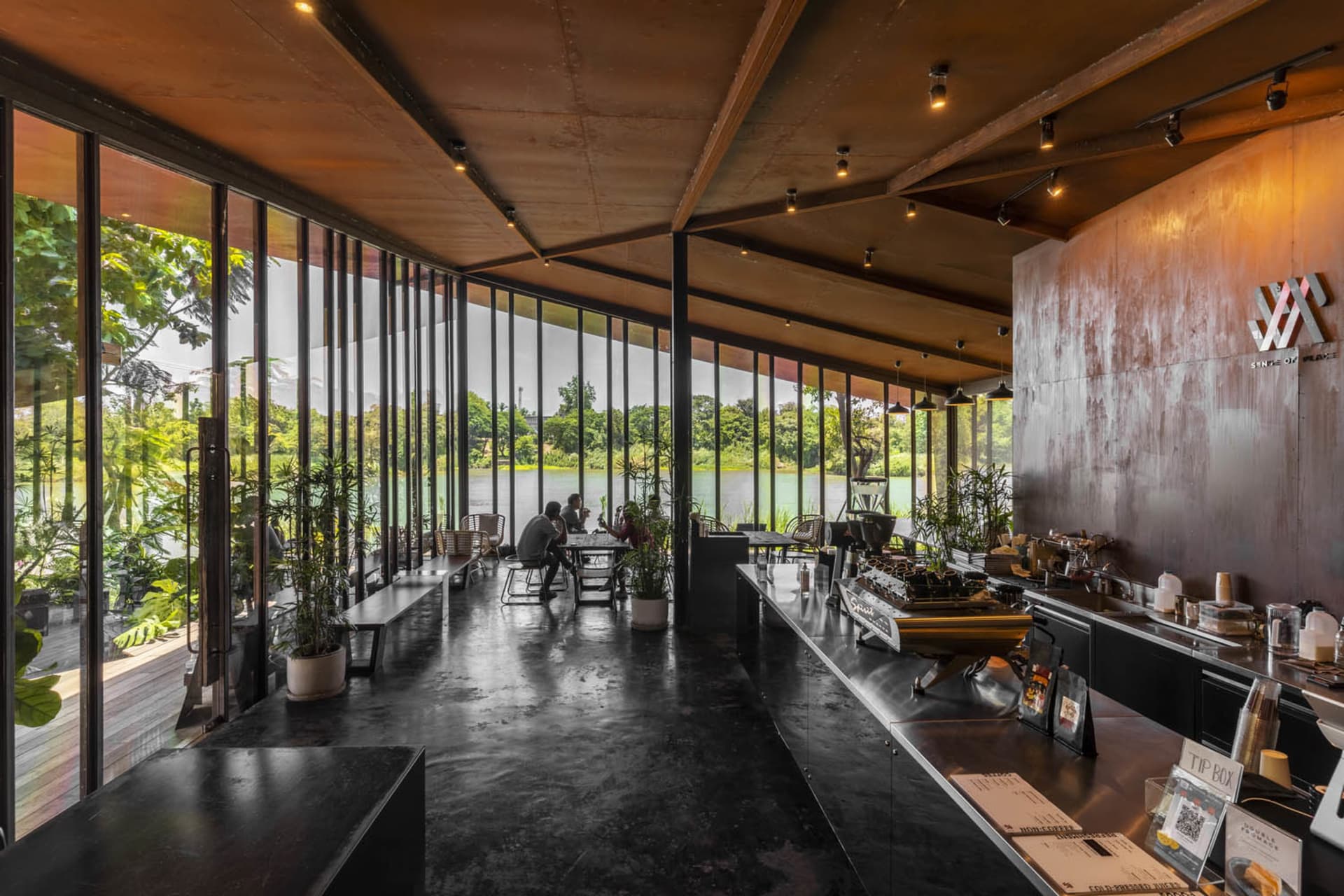 A bright and welcoming cafe with dining tables, chairs, potted plants, and cups and bowls scattered around, set against a backdrop of a large window and natural light