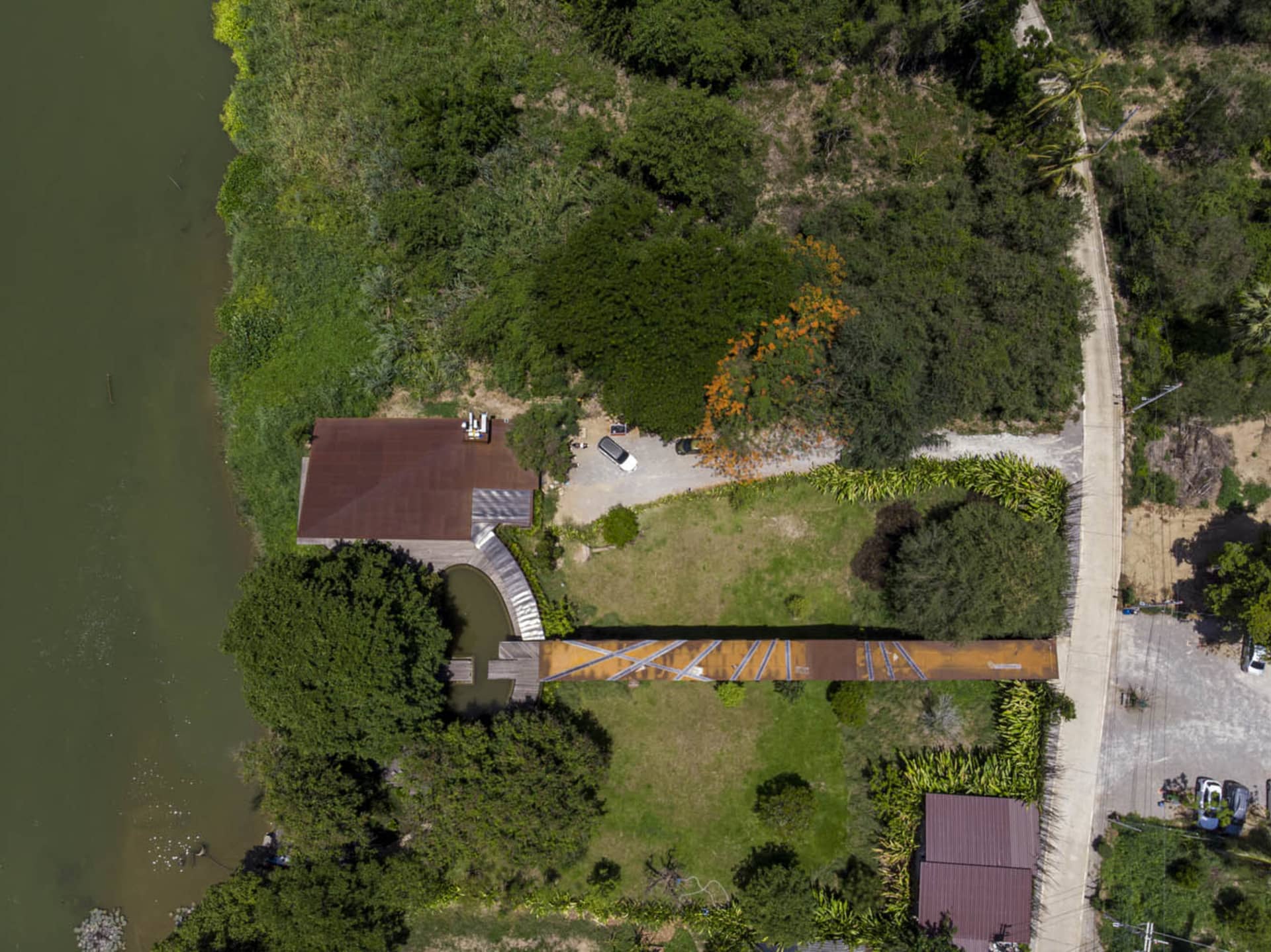 A house with a brown roof and a wooden bridge overlooking a river, surrounded by trees and a parked car