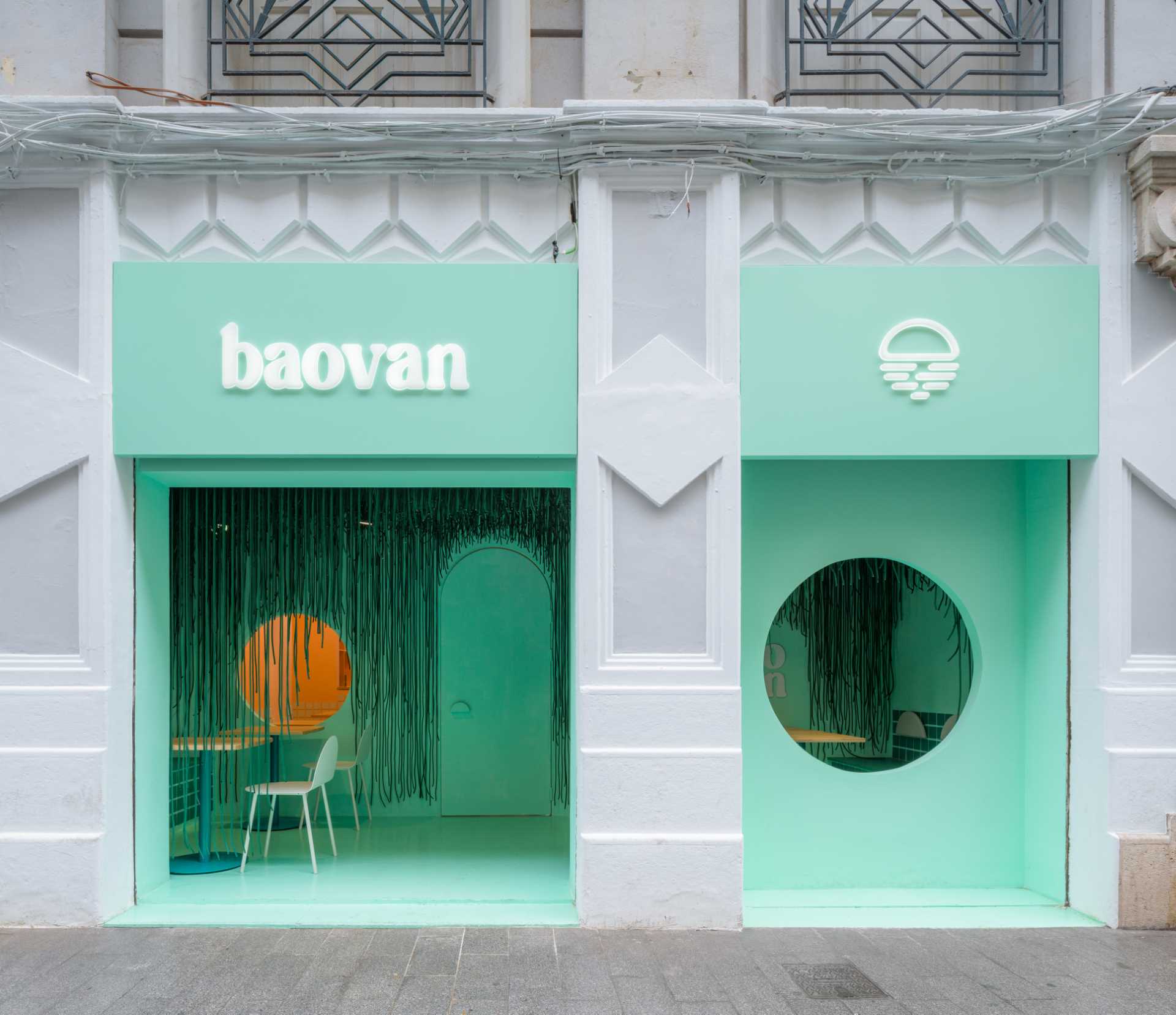 Two modern storefronts with green walls and white trim, one with a circular window and the other with a doorway, surrounded by chairs and a dining table in a well-lit commercial space