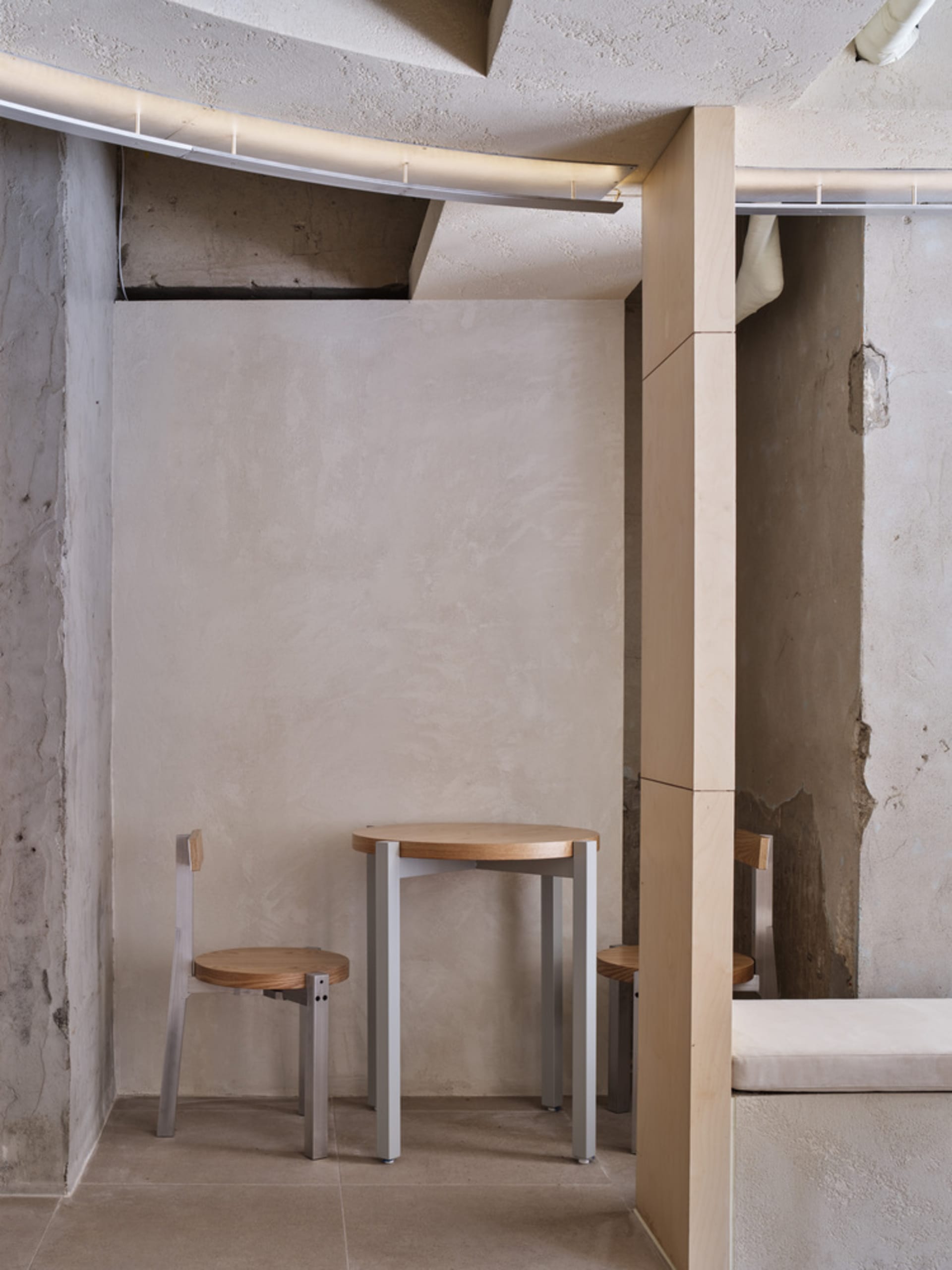 Dining area with concrete walls, a small table, and three chairs, accompanied by a bench in a modern and industrial space