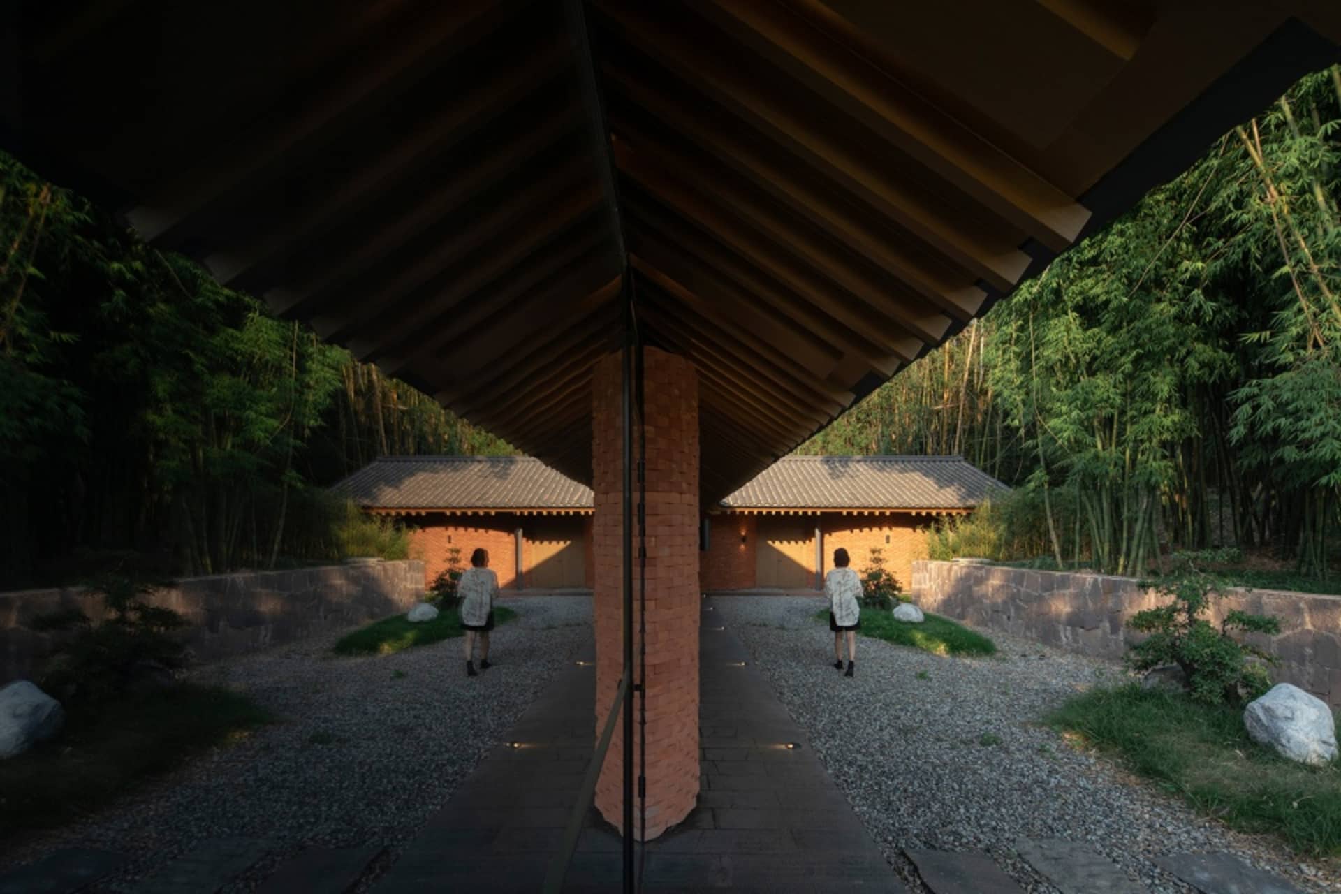 A woman walks towards the entrance of a traditional Asian building with a brick wall, wooden roof, and bamboo elements