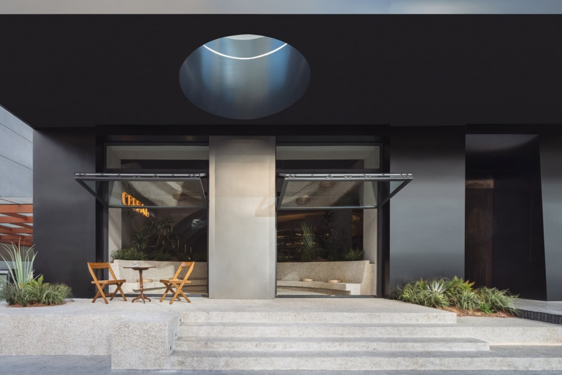 A modern building exterior with a black facade, a large circular window, and two chairs outside