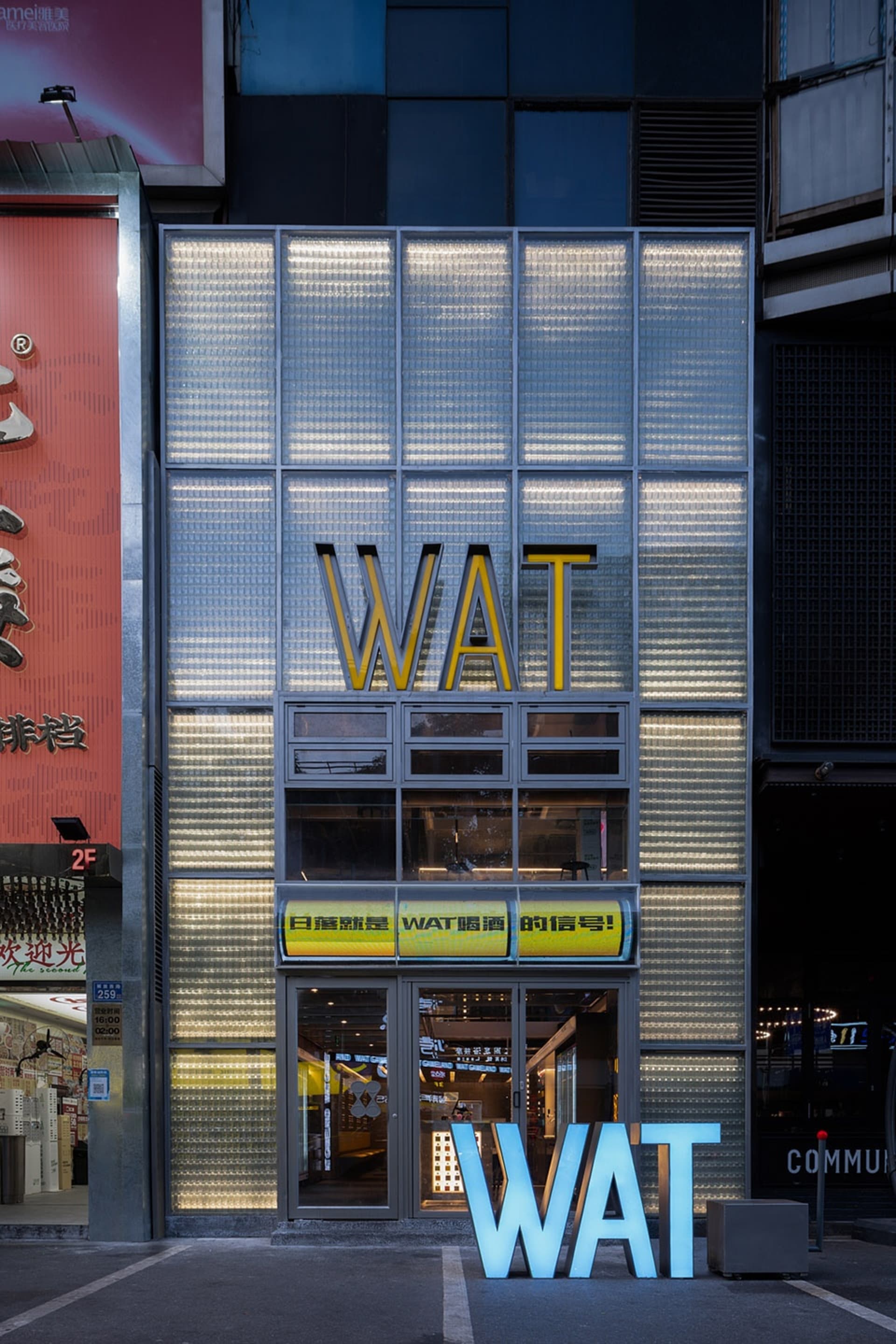 A bustling Asian city street with two large buildings, one with a yellow sign reading WAT and the other with a red banner featuring Chinese characters, surrounded by parked cars and a lively urban atmosphere
