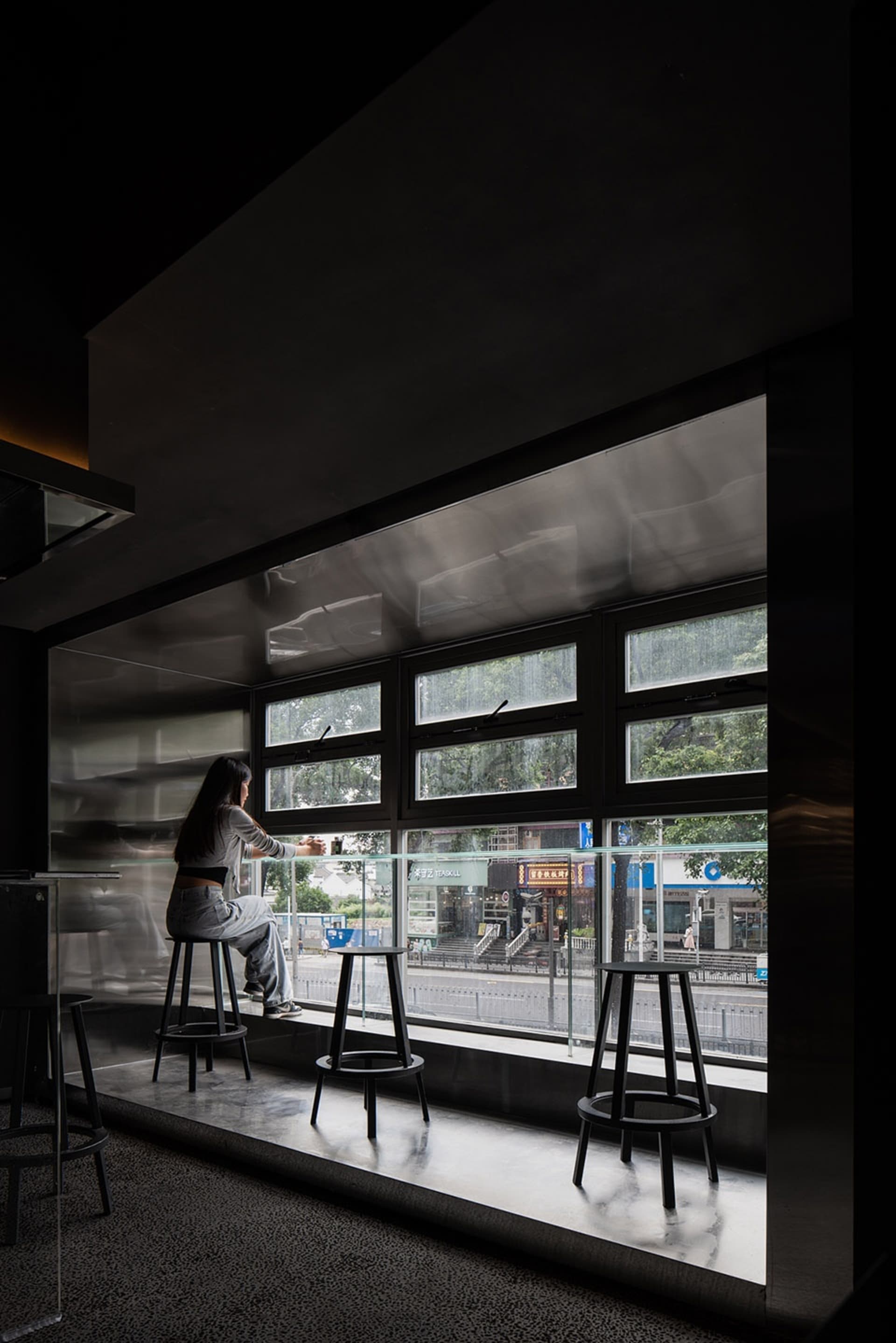 A woman sits at a counter in a cafe with a glass window, surrounded by chairs and a dining table, in a calm and relaxed atmosphere