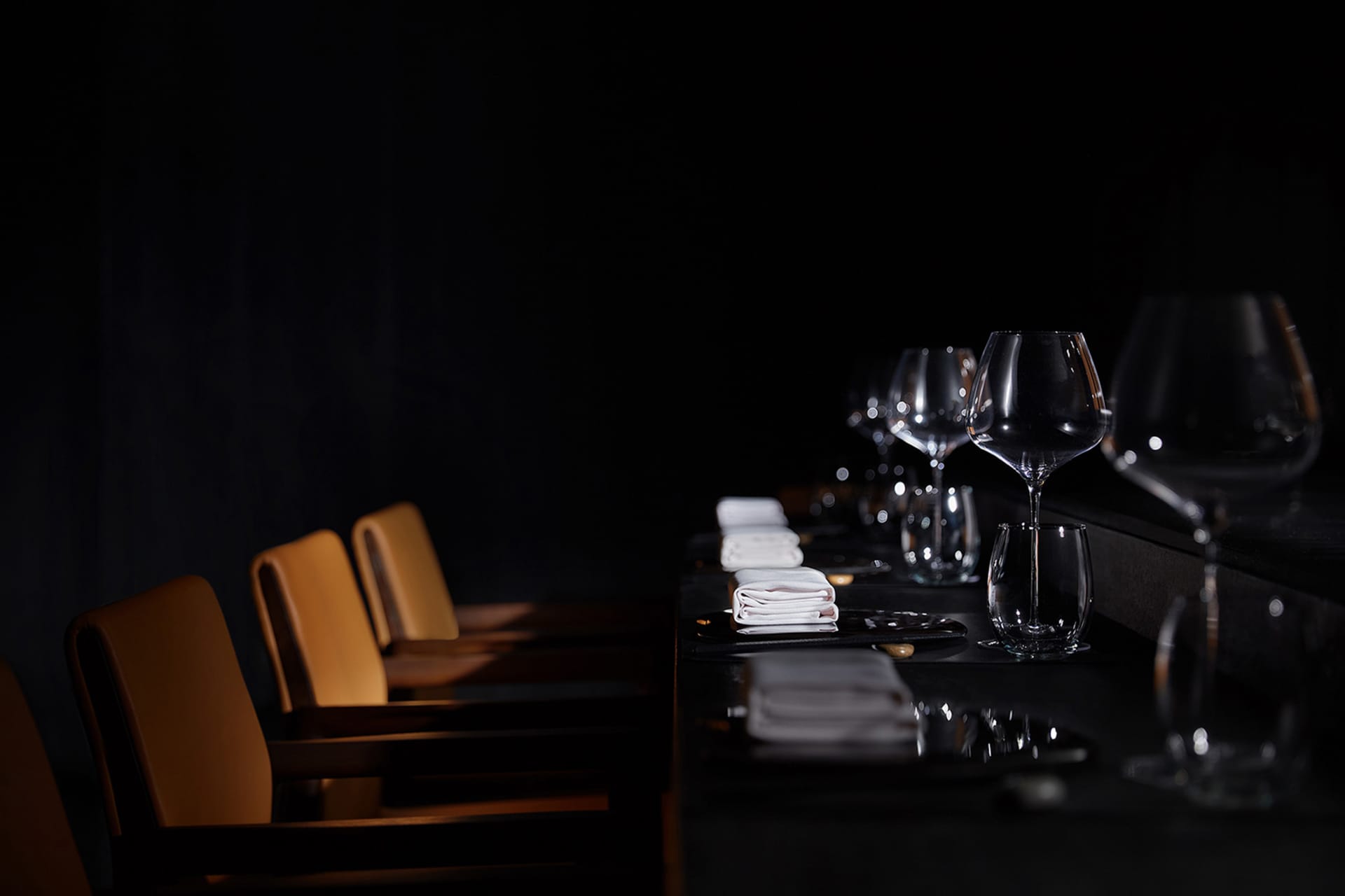A darkened dining room with a wooden table and chairs, wine glasses, and napkins, creating a sophisticated atmosphere