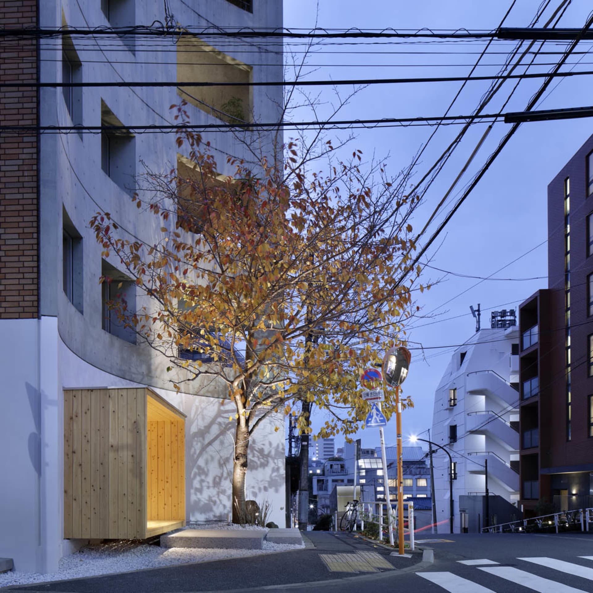 A city street scene with a tall building, a tree with orange leaves, two traffic lights, and three chairs in a seating arrangement