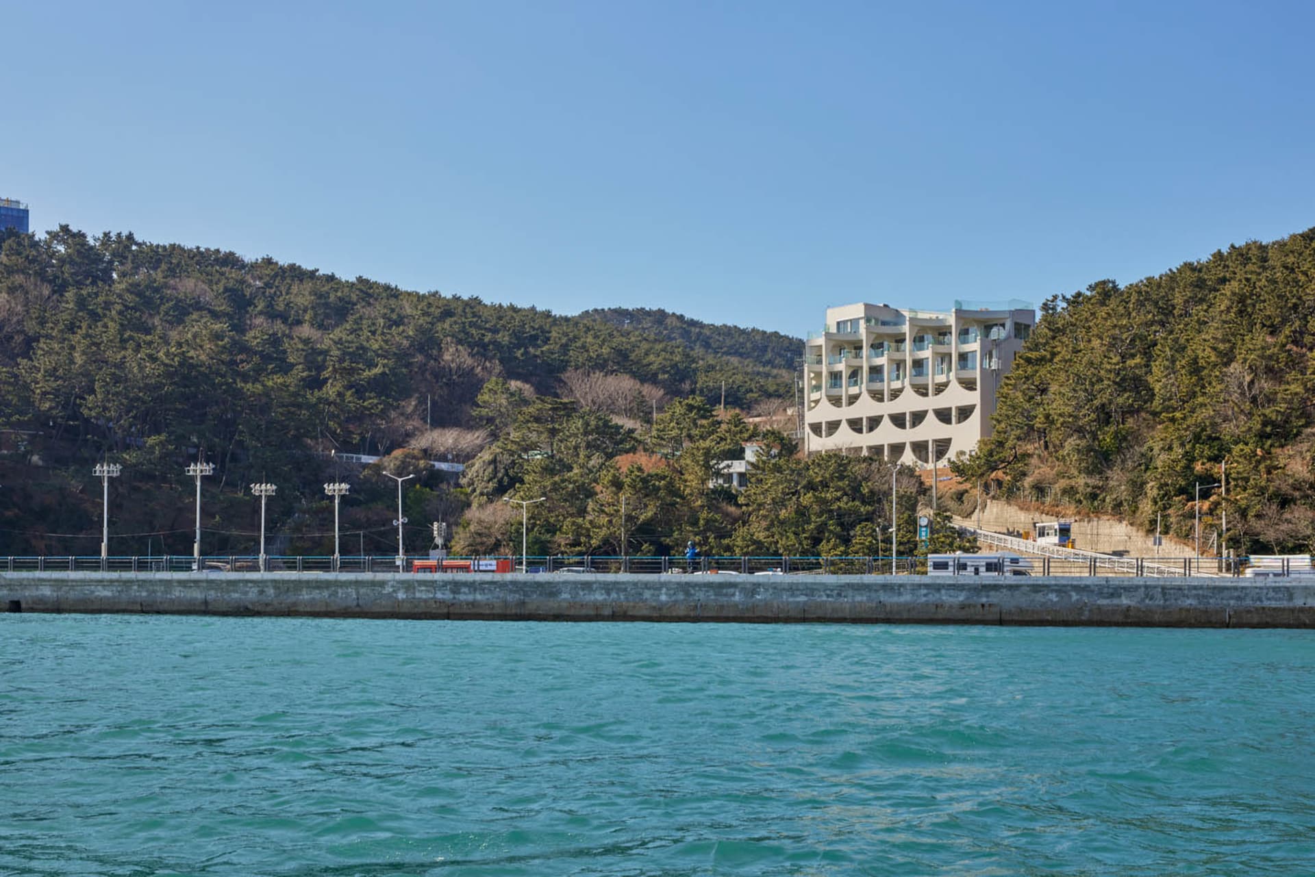 A modern building with a concrete facade situated on a hillside overlooking a body of water, with several cars parked in front