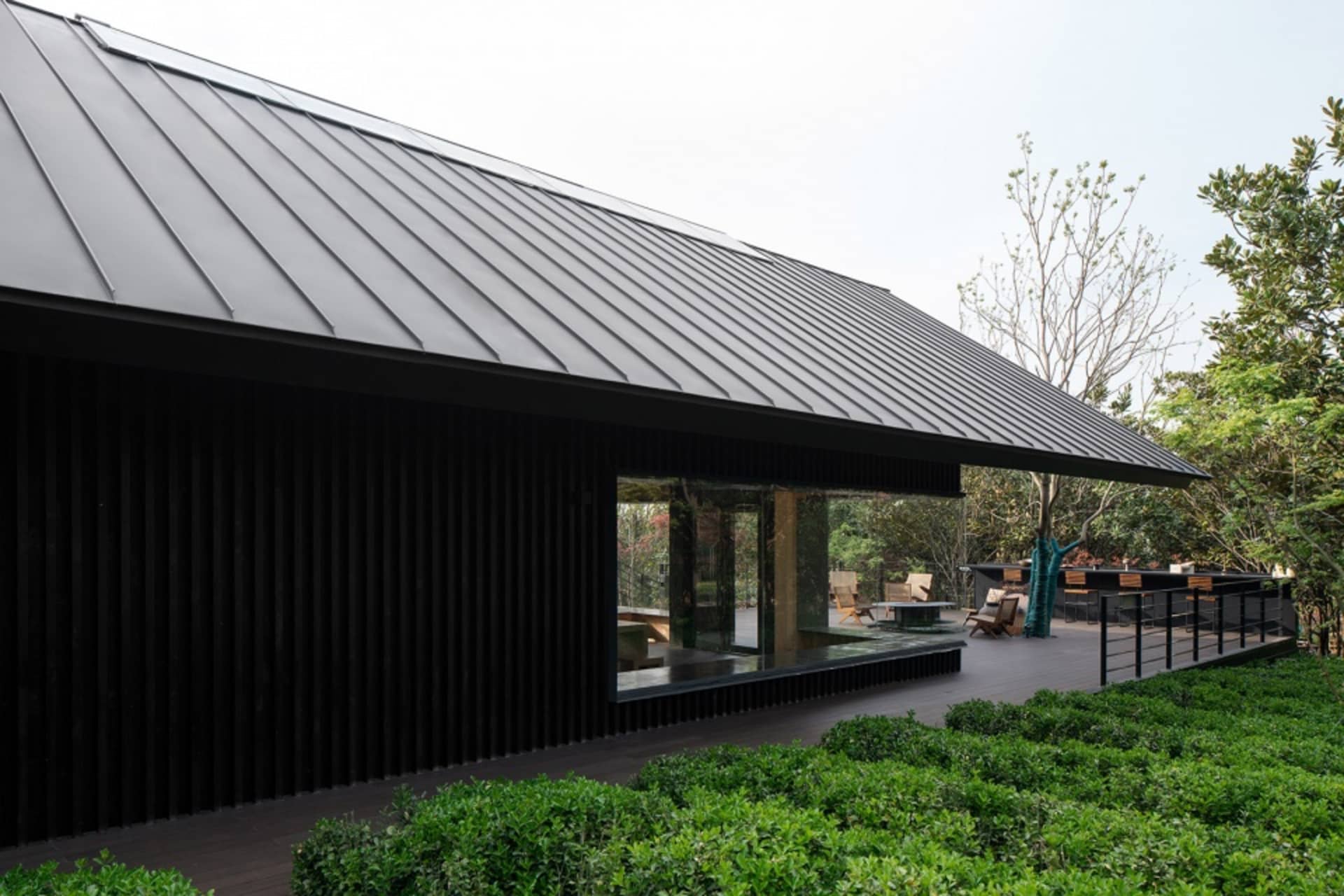 A modern house with a black roof and green facade features a large window and outdoor seating area with chairs and a bench, set in a calm and inviting atmosphere